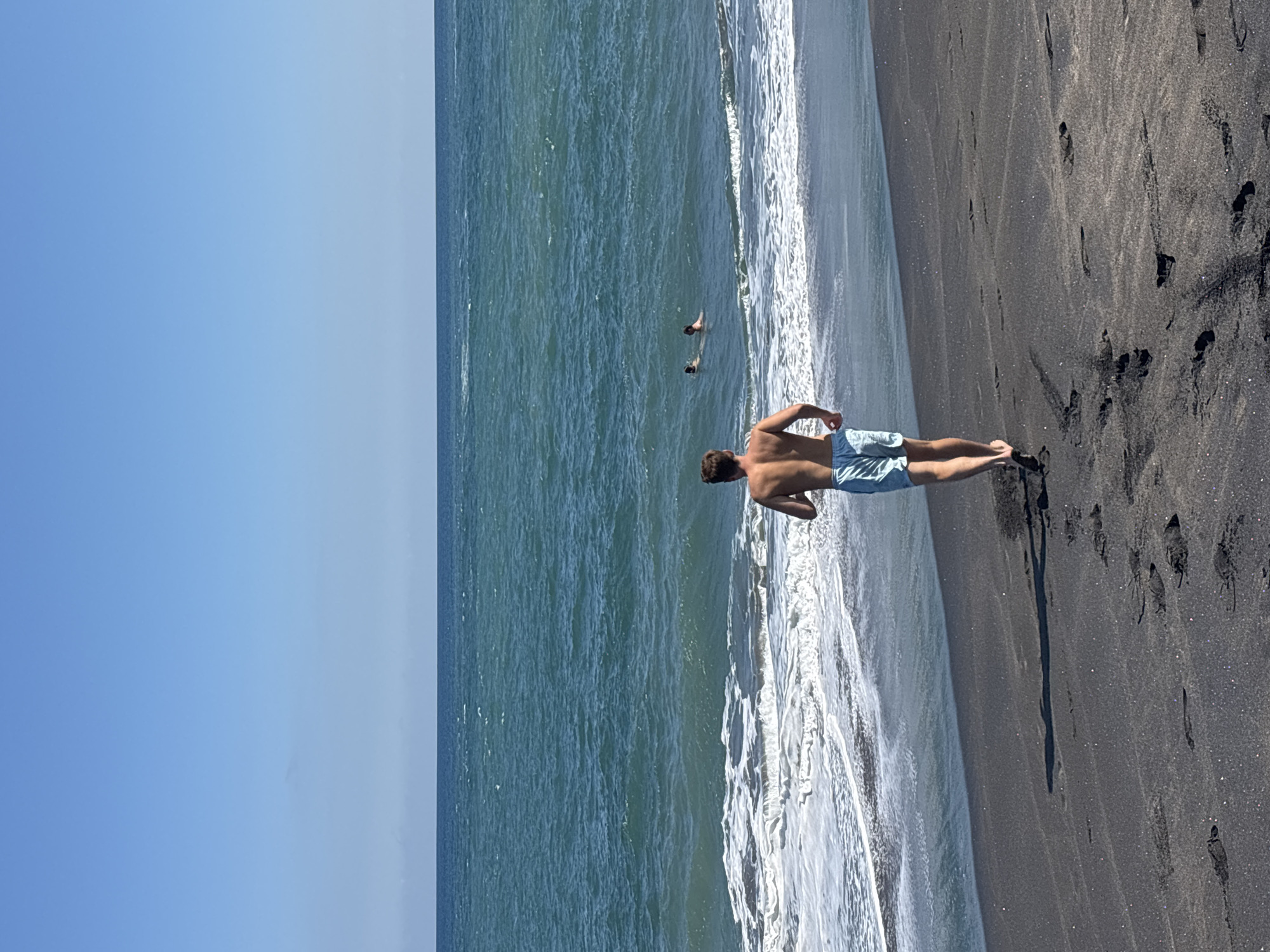A man is running on a black sand beach towards the ocean, which has moderate waves and a clear blue sky above. In the distance, two people are swimming in the water. The man is wearing light blue swim trunks. The sand is wet and dark, with footprints visible. The location is the beach in Mengwi, Indonesia.