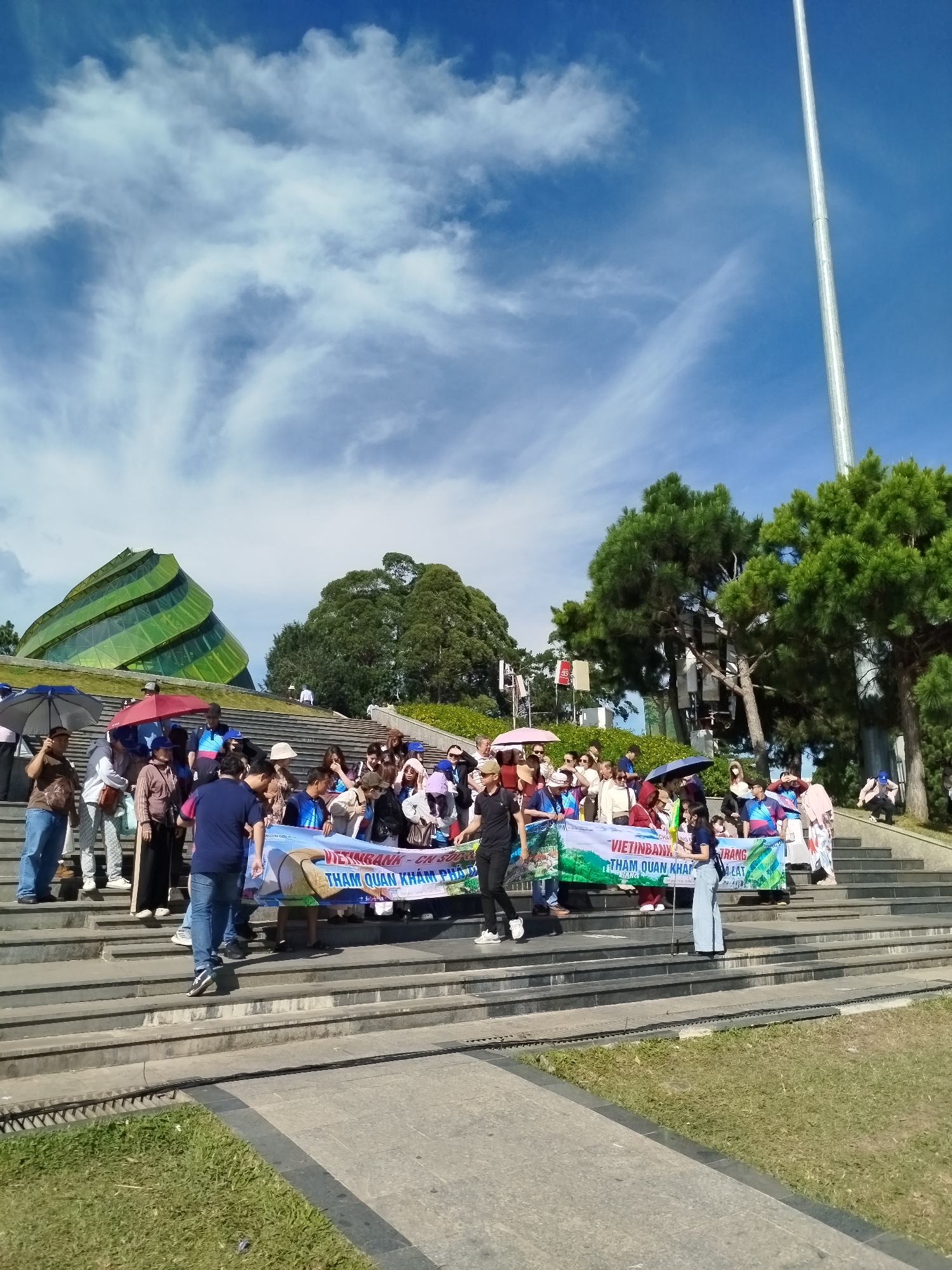 A large group of people are gathered on wide stone steps under a partly cloudy blue sky. Many people are holding umbrellas, some pink, some blue, and one grey. A long banner is held across the front of the group, displaying text in Vietnamese. The banner reads "VIETINBANK - CN SUOI VANG - CHUNG TAY KHAM PHA DA LAT" which translates to "VIETINBANK - Suoi Vang Branch - Joining Hands to Explore Da Lat".
In the background, to the left of the frame, is a distinctive green building with a curved, spiral design. Behind the people and to the right are lush green pine trees. A tall, thin, metallic pole extends upwards from behind the trees and into the sky. The steps lead up a grassy slope. A paved walkway with a strip of grass runs along the bottom of the image. The scene appears to be a tourist attraction.