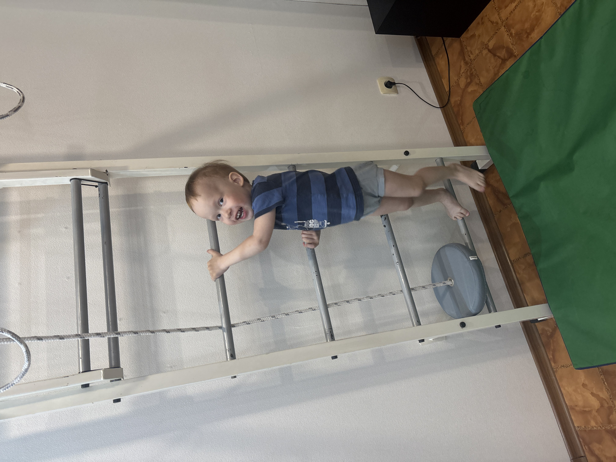 A child is shown lying horizontally on a gymnastic wall bar set in a room. The wall bars are white with grey rungs. The child is wearing a navy and blue striped shirt and grey shorts. The child is holding onto a rung with each hand and facing the camera with an open mouth and slightly wild look. The background consists of a beige wall with a black speaker cabinet in the top right corner. A power outlet with a plugged-in cord is visible on the wall. A green mat is partially visible on the floor in the top right. The floor is tiled in a brown and tan pattern. The location is identified as Ферма, Russia.