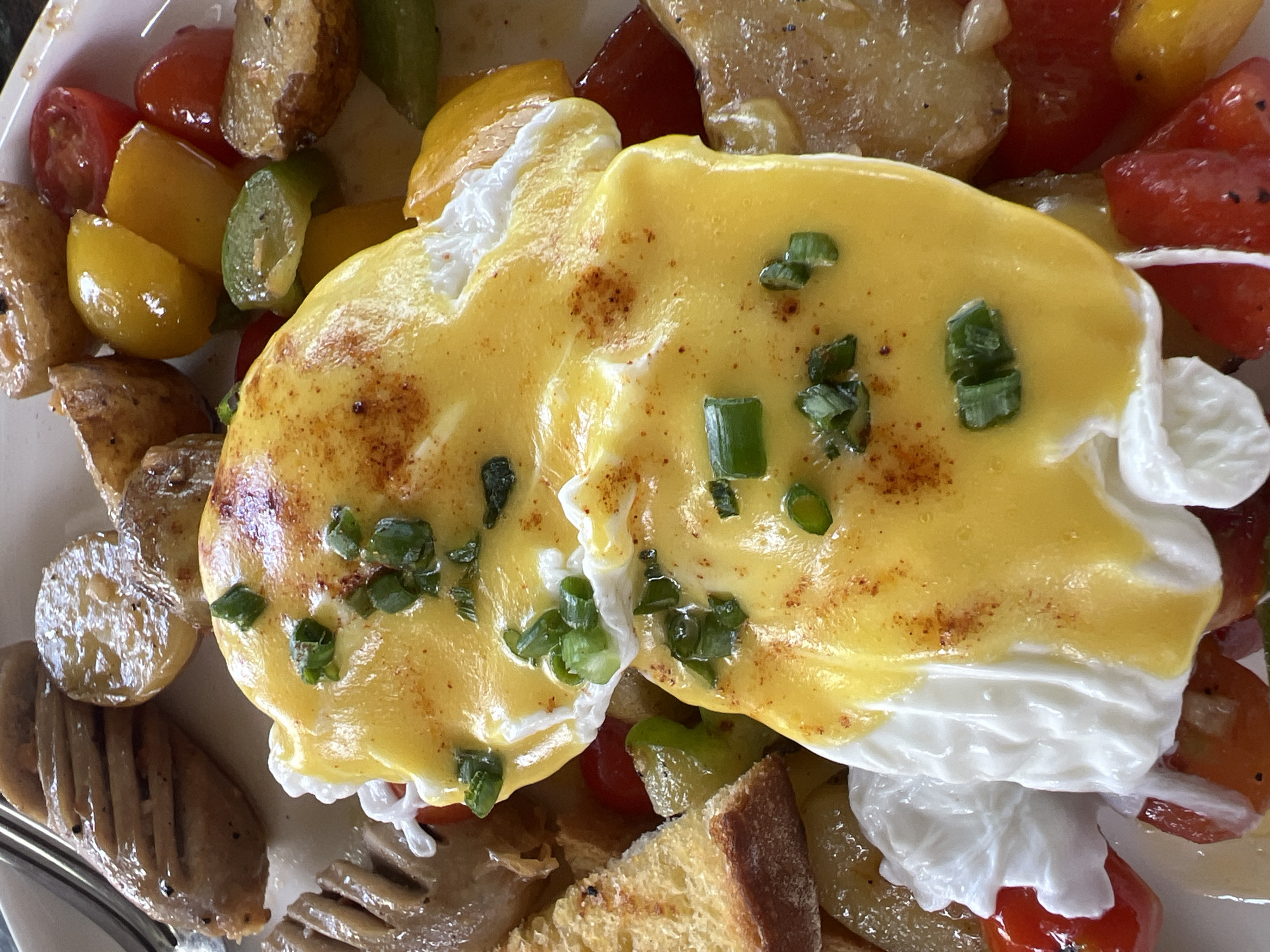 This close-up image captures a vibrant and hearty breakfast or brunch dish served on a white plate. The main focus is two perfectly poached eggs, generously covered in a rich, bright yellow hollandaise sauce. These are artfully garnished with finely chopped green onions or chives and a light dusting of reddish spice, possibly paprika.
Surrounding the eggs is an assortment of accompanying ingredients. These include roasted or pan-fried potato pieces, some still retaining their skins, and a colorful mix of halved cherry tomatoes in both red and yellow hues, along with chopped green and yellow bell peppers. Towards the lower left of the frame, several slices of a grilled, sausage-like item are visible, showing distinct char marks from the grill. A piece of toasted bread or a crouton is also present near the bottom right.
The food is brightly lit, highlighting the fresh colors and textures. The meal appears well-prepared and is typical of a substantial breakfast or brunch offering from a restaurant or cafe in North Kuta, Indonesia. No text is visible within the image.