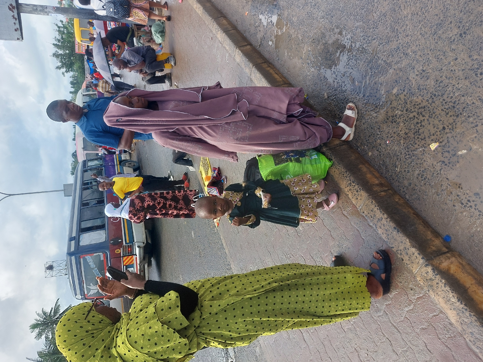 The image depicts a street scene in what is likely Dar es-Salaam, Tanzania, given the visible buses and general environment. A woman in a green polka-dot dress and head covering stands near the curb of a street, holding a black object, possibly a phone. Beside her stands a young child in a patterned dress and a dark-colored jacket. Further along the curb, a man wearing a blue shirt is seated, holding a woman wrapped in a purple shawl or cloth. Several other people are visible in the background near what appears to be a bus stop, including individuals standing, sitting on stools, and carrying umbrellas. Buses of varying colors are also parked in the background, with one featuring visible text in Swahili, the national language of Tanzania. The street surface near the curb shows signs of wetness, possibly from recent rain.