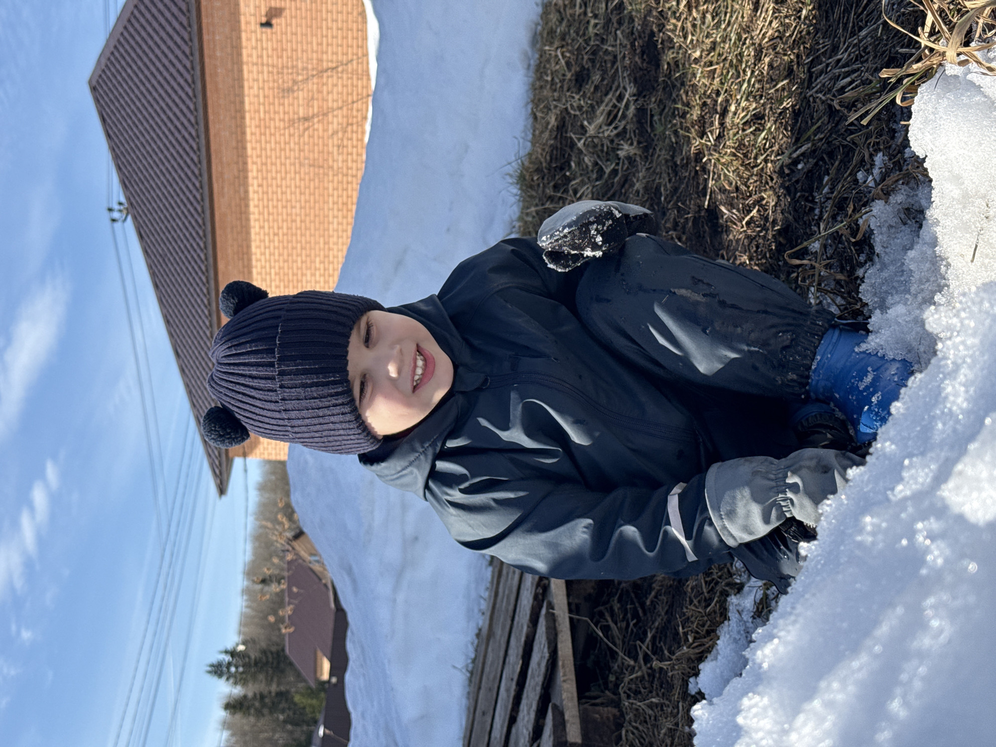 A young child is depicted crouching outdoors in a partially snow-covered residential area of Chaikovsky, Russia, under a clear blue sky. The child, approximately 4-6 years old, wears a dark blue ribbed knit beanie with two pom-poms resembling animal ears, a dark blue hooded waterproof snowsuit featuring a reflective stripe on the left forearm, grey insulated gloves, and blue boots. The child's face is smiling, with teeth visible, and directed downwards towards the ground. The child's left gloved hand is submerged in a small bank of granular snow in the immediate foreground. The ground consists of patches of melting snow, exposed brown grass, and dark soil. To the child's left, a stack of worn wooden pallets rests on the ground. A substantial snowdrift extends horizontally across the mid-ground. Behind this snowdrift, to the right, stands a two-story house constructed of orange-brown brick with a dark brown tiled gabled roof. Further in the background, another smaller building with a dark roof is partially visible among bare deciduous trees and dark green coniferous trees. Multiple overhead power lines traverse the upper portion of the clear blue sky.