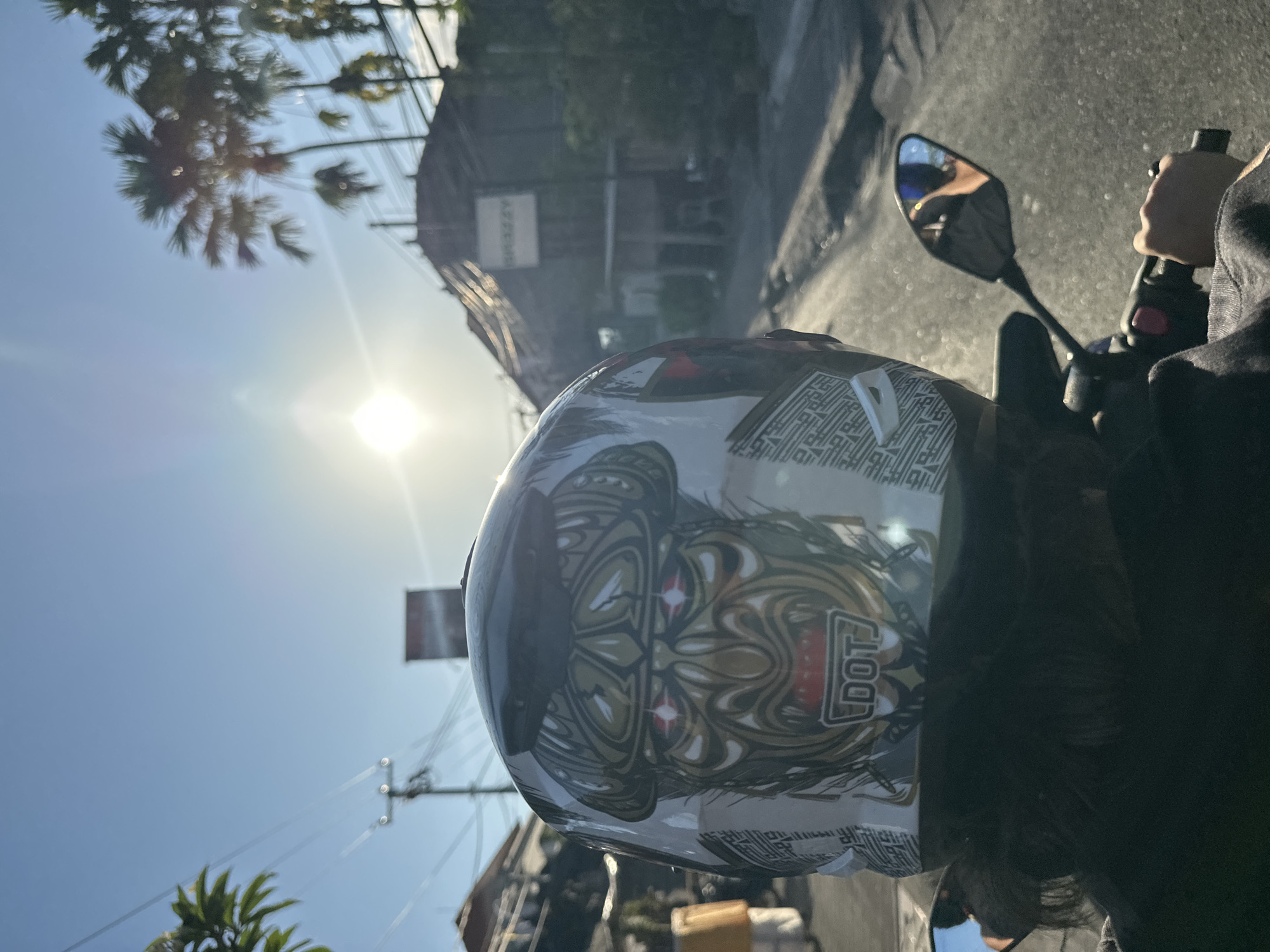 The image, taken from a first-person perspective, shows a person riding a motorcycle in North Kuta, Indonesia. The rider is wearing a motorcycle helmet with intricate designs and the letters "DOT" visible. A side mirror and handlebar are partially visible. The background reveals a bright, sunny day with palm trees, buildings, and street infrastructure, partially obscured by glare. The road ahead appears wet or reflective.