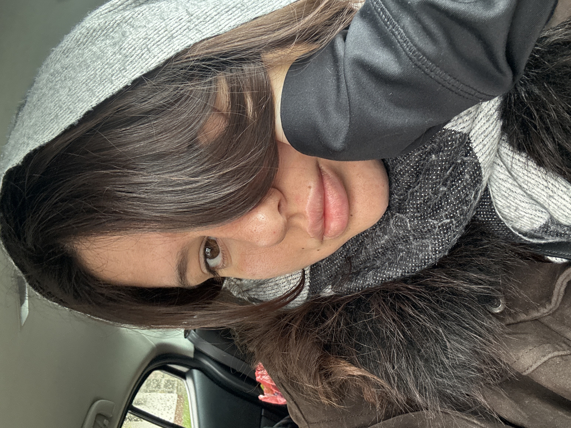 A woman is sitting inside a car. She is wearing a grey hooded sweatshirt, a black long-sleeved shirt, and a grey patterned scarf. She appears to be wearing a brown jacket with a fur collar. Her brown hair is partially obscuring her face. The scene is framed by the car's interior, with a partial view of the outside through the car window. Based on the available visual information, it's impossible to determine if the photo was taken in Burgas, Bulgaria.