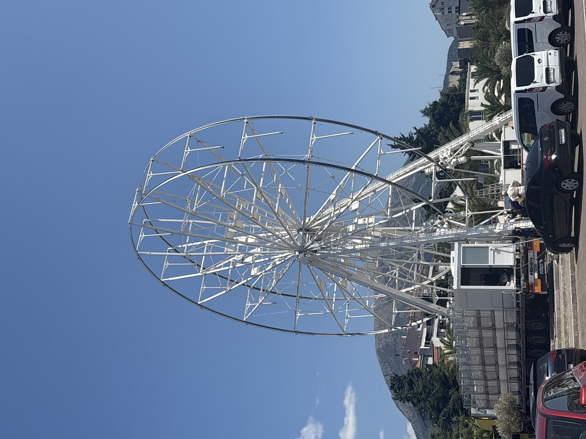 This image shows a large, white Ferris wheel under construction in a city. The Ferris wheel is the dominant feature, its metal framework stark against a clear blue sky. In the foreground, several vehicles are parked, including a red car, a dark sedan, and three white vans. To the left of the Ferris wheel, a large truck with a trailer filled with stacked metal cages is visible. Workers can be seen near the truck and the base of the Ferris wheel, suggesting assembly or maintenance activities. In the background, buildings and lush green trees, including palm trees, are present, with a mountain range visible behind them. The overall scene appears to be outdoors in a sunny, urban environment. Given the visible signage on the truck, "SCHMITZ" and what appears to be a license plate from Montenegro, this scene is likely in the city of Bar, Montenegro.