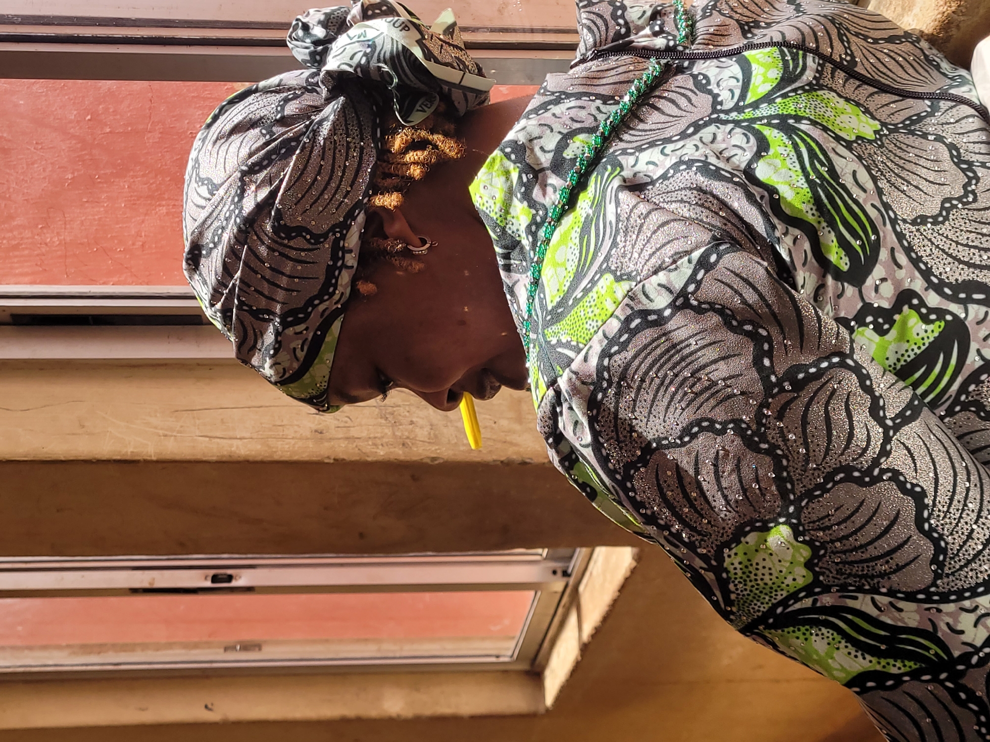 A woman wearing a patterned headwrap and matching top is bent forward, looking downward. She has golden hair decorations and jewelry in her ear. She has a yellow object in her mouth. Behind her are horizontal wood beams and a window with a reddish hue. The location is implied to be Bukuru, Nigeria.