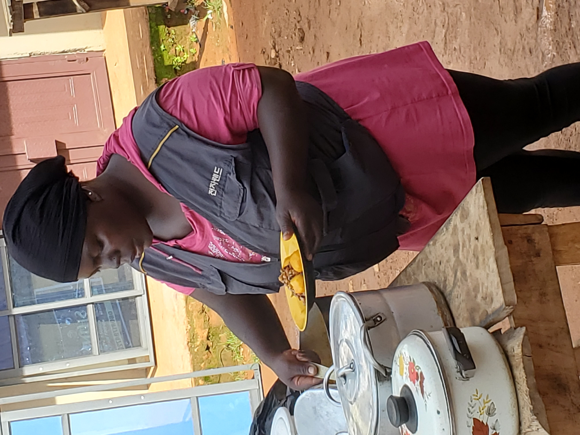 The media file captures a person in Bukuru, Nigeria, engaged in food preparation or serving. The subject, who appears to be a woman wearing a pink shirt and a dark apron with yellow trim, has dark skin and is wearing a black headscarf. She is holding a yellow plate with food, possibly plantains and beans, and appears to be in the process of serving it. In front of her are several cooking pots with lids, some of which are decorated with floral patterns. The setting suggests an outdoor or semi-outdoor environment, with a building and possibly a window visible in the background, along with a dirt ground and some greenery. The light suggests it is daytime. Visible text on the apron reads "전자랜드" in Korean, which translates to "Electronics Land" and is a Korean electronics retailer.