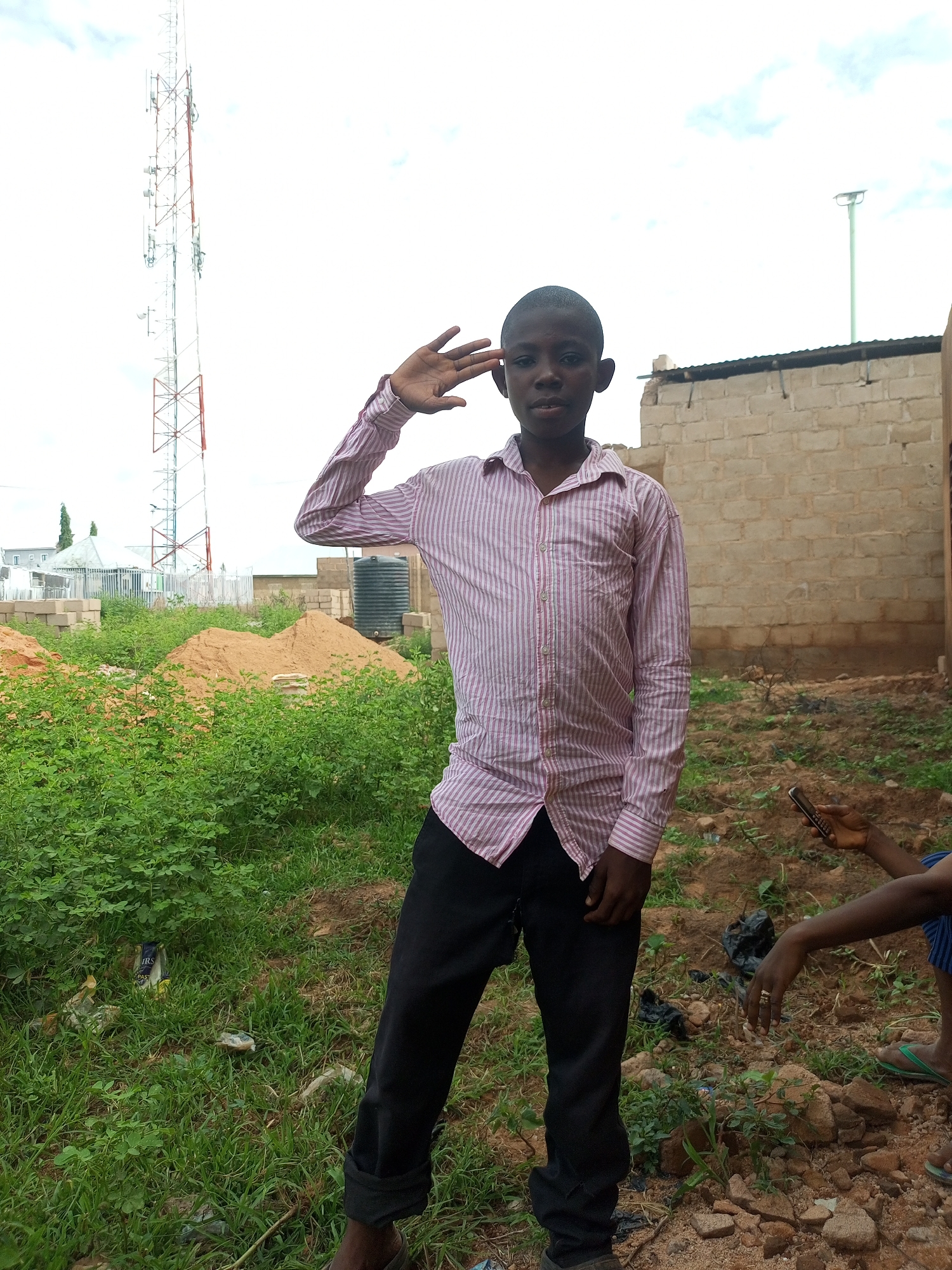 A young boy, appearing to be of African descent, stands in the foreground, facing the camera. He is wearing a pink and white striped button-down shirt and black pants. His right hand is raised to his forehead in what appears to be a salute. Behind him, to the left, is a tall telecommunications tower. To the right, there is a brick building under construction. In the mid-ground, there are piles of sand and some dense green vegetation. The ground is a mix of dirt and small rocks. The sky is overcast. The scene appears to be an outdoor area within the city of Tempure, Nigeria, likely a construction site or an undeveloped area.