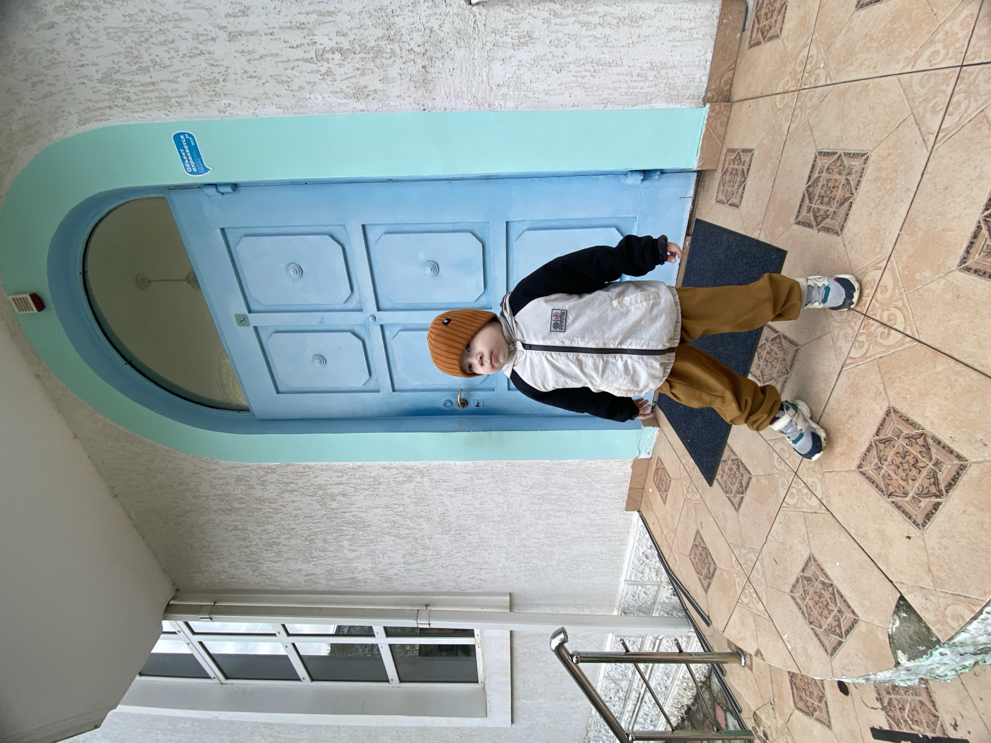 A child wearing a brown beanie, a white and black jacket, and brown pants stands on tiled flooring in front of a blue door with a curved blue frame. The door is framed by a white wall. There is a window below the wall and a metal railing. The flooring is tiled with patterned beige tiles and a small black mat. A sign with Cyrillic writing is on the blue door frame. The location is most likely Sochi, Russia.