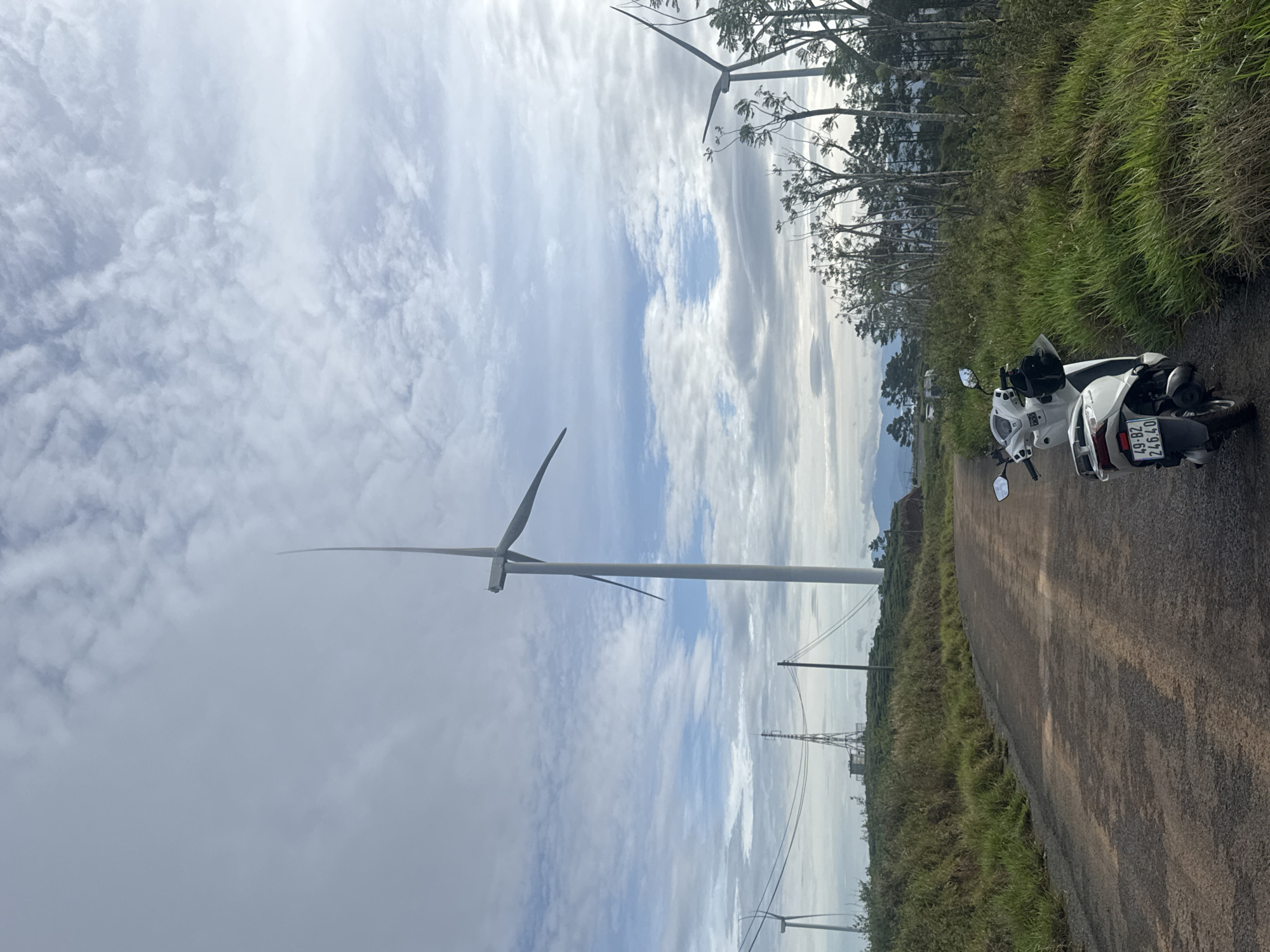 A white scooter is parked on the right side of a paved road. The road stretches into the distance, where wind turbines are visible on both sides. The surrounding landscape consists of grassy hills and trees. Above, the sky is filled with clouds. The scooter's license plate appears to read "49-B2 246.40". The setting is likely in the vicinity of the Trường Thọ wind farm, Vietnam.