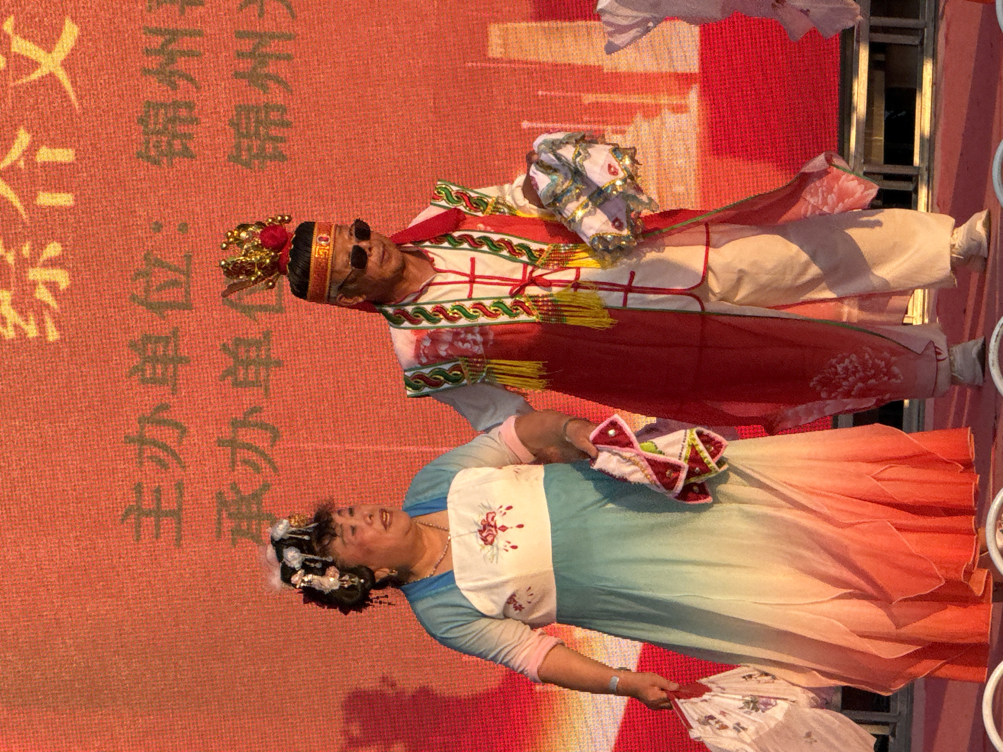 In Jinzhou, China, a man and a woman, dressed in elaborate traditional attire, stand on a stage. The woman wears a layered, gradient dress of blue to orange, adorned with intricate floral embroidery on the bodice, and a decorative headpiece. She holds a folded white fan in her left hand and a patterned cloth in her right. The man, wearing sunglasses, sports a red and white ensemble with ornate trim, a golden crown-like headdress, and holds a similar patterned cloth. Behind them, a red backdrop displays Chinese characters, likely indicating sponsors or event details. The lighting suggests an outdoor evening event, possibly a cultural performance or festival.