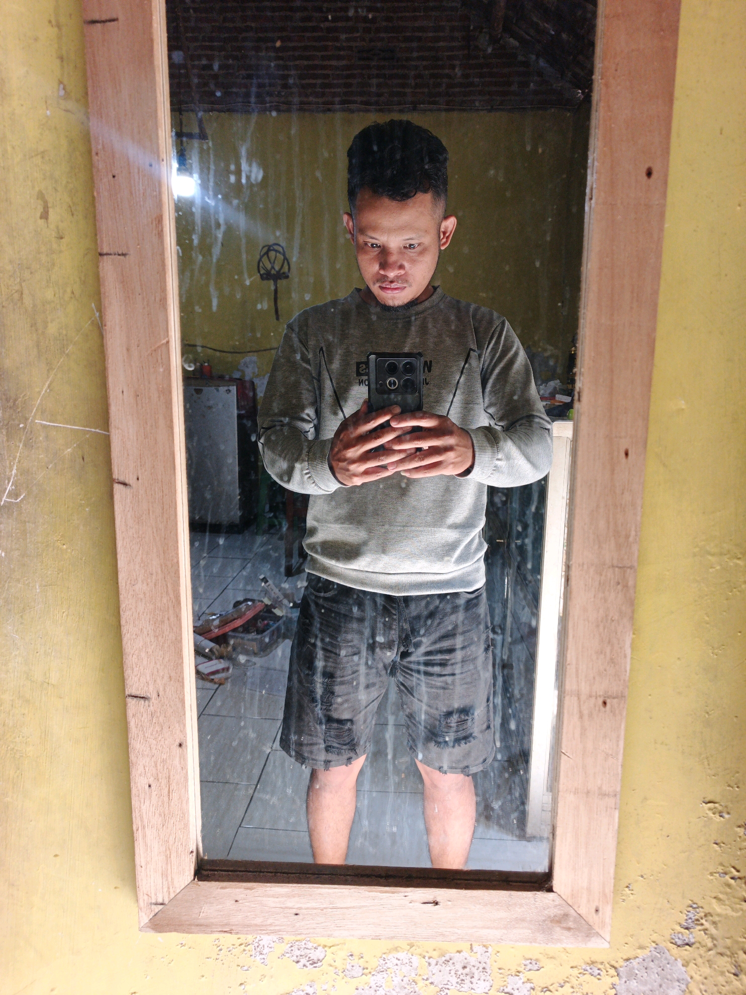 A full-length mirror, framed with rough-hewn wood, reflects a young man standing in what appears to be a storage area or a workshop in Modongan, Indonesia. The man, with short, dark, curly hair and wearing a grey long-sleeved shirt and dark denim shorts, holds a black smartphone in his right hand, taking a selfie. His face is centered in the mirror, with his gaze directed downwards at the phone screen.
The background reflected in the mirror reveals a cluttered environment. Yellow walls are visible, with what looks like whitewash or paint peeling in places, suggesting an older or unfinished structure. The floor is tiled. Various objects are scattered in the reflection, including a metal cabinet, what could be tools or supplies in containers, and a bare light bulb hanging from the ceiling. The overall impression is one of a utilitarian space, possibly a garage or an indoor workspace, rather than a living area. The wooden frame of the mirror itself shows signs of construction, with visible nails. The lighting is dim, with the primary light source appearing to be the overhead bulb reflected in the mirror.