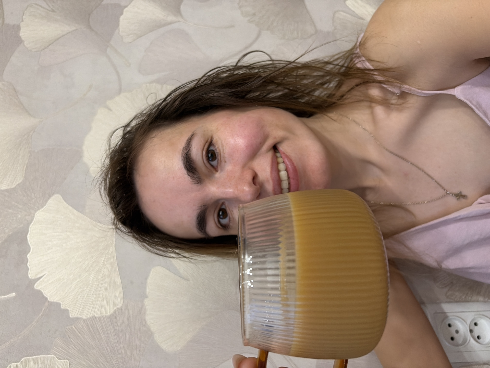 A woman with wet brown hair is holding a clear glass mug filled with a light-brown beverage, possibly coffee with milk, up to her face and smiling at the camera. She is wearing a pink tank top and a silver necklace with a small cross pendant. The background is a wall with a floral pattern, specifically resembling ginkgo leaves. An electrical outlet is visible in the lower right corner of the frame. The image orientation is sideways. There is no information to confirm that this image was taken in Izhevsk, Russia.