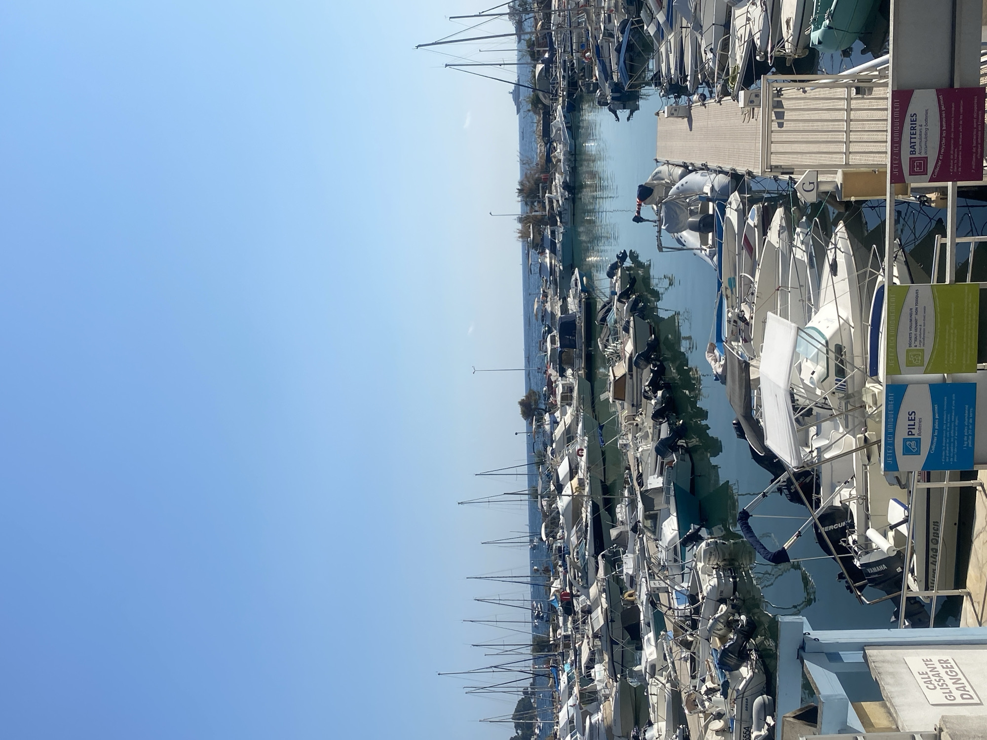 This is an exterior, eye-level, vertical, medium shot of a harbor in Antibes, France, on a clear, sunny day. The sky is a bright, cloudless blue, and the water in the harbor is a calm, reflective blue. The harbor is filled with numerous boats of various sizes, mostly small to medium-sized pleasure crafts. Many of these boats are sailboats, identifiable by their tall masts. There are also motorboats and what appear to be rigid inflatable boats. The boats are docked in neat rows.
In the foreground, to the left, is a blue structure with a sign that reads "CALE GLISSANTE DANGER" (Slipping Ramp Danger). Further to the right, there are several signs indicating waste disposal for "PILES BATTERIES" (Batteries) and "DECHETS VOLUMINEUX A "TOUT VENANT" NON TOXIQUES" (Bulky Waste of "Any Kind" Non-Toxic). A wooden walkway extends into the harbor, with several boats docked alongside it.
In the background, more boats are visible, and beyond the harbor, there is a shoreline with vegetation and buildings. The sea extends to the horizon, with a few distant boats and a larger vessel resembling a yacht visible in the far distance. The overall impression is of a bustling, well-organized marina on a beautiful day. No people are clearly visible in the scene.