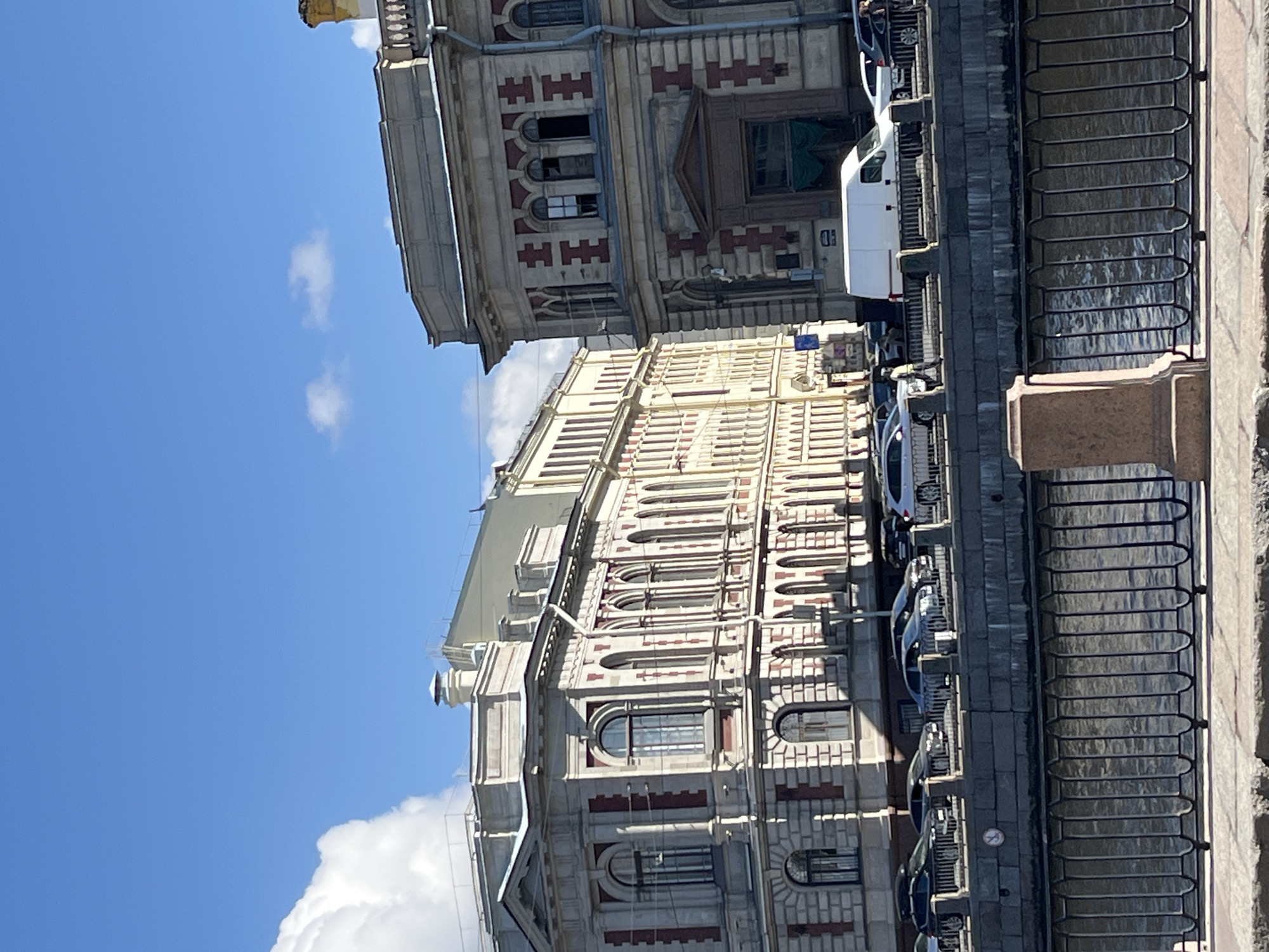 The image shows two ornate buildings in Saint Petersburg, Russia, under a blue sky with white clouds. The buildings are adorned with architectural details such as arched windows, cornices, and decorative brickwork. A wrought iron fence runs along the edge of the waterfront. Cars and a white van are parked along the street adjacent to the buildings. The water is visible behind the fence.