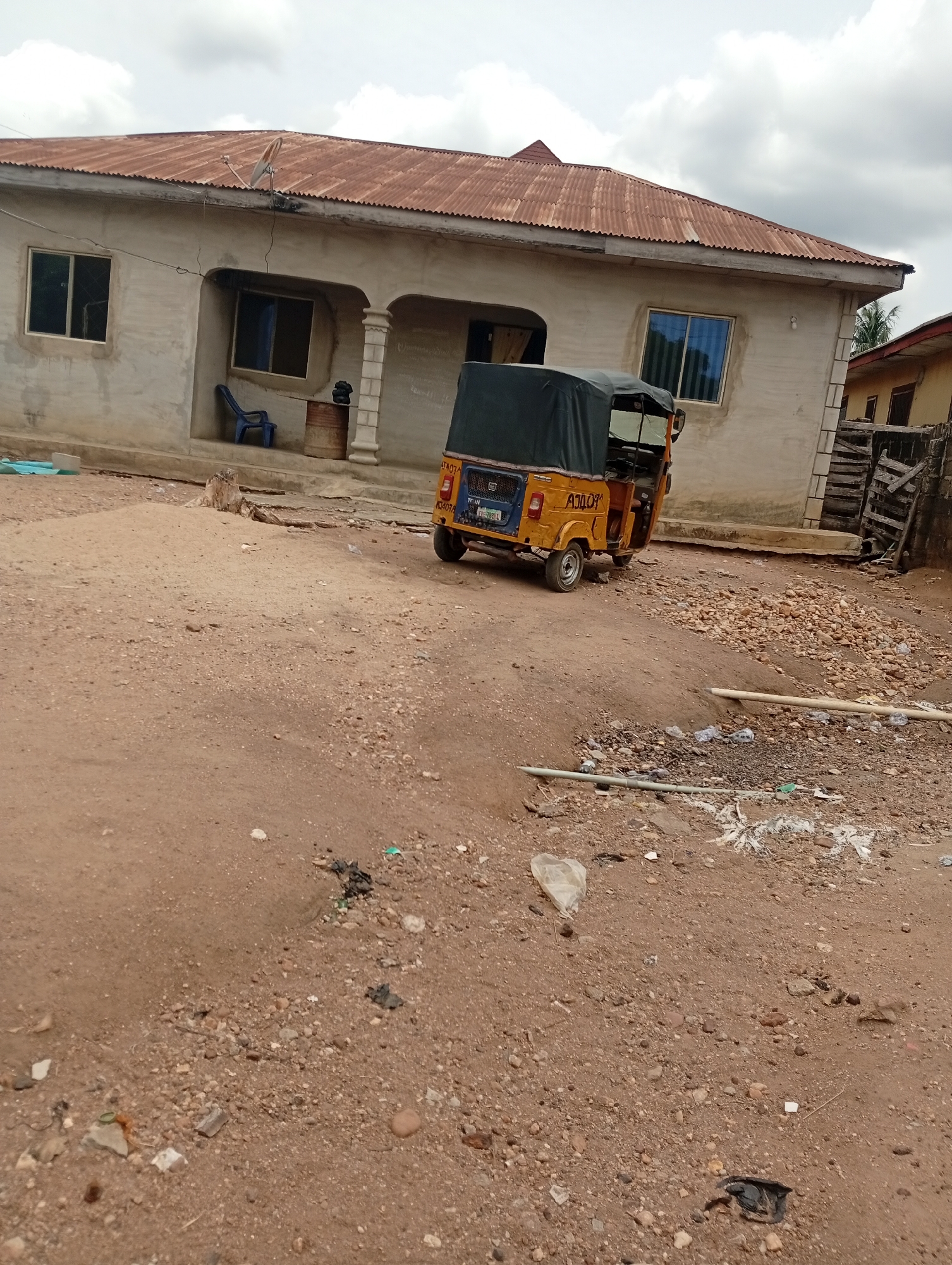The image depicts a scene in what appears to be a residential area in Adankolo, Nigeria. In the background is a one-story building with a light-colored exterior and a corrugated metal roof. The building has several windows, and a small porch area supported by columns. Parked in front of the building is a yellow "Keke" or auto-rickshaw, a common mode of transportation in Nigeria. The foreground shows a dirt ground littered with small stones, debris, and plastic waste. No people are visible in the image.