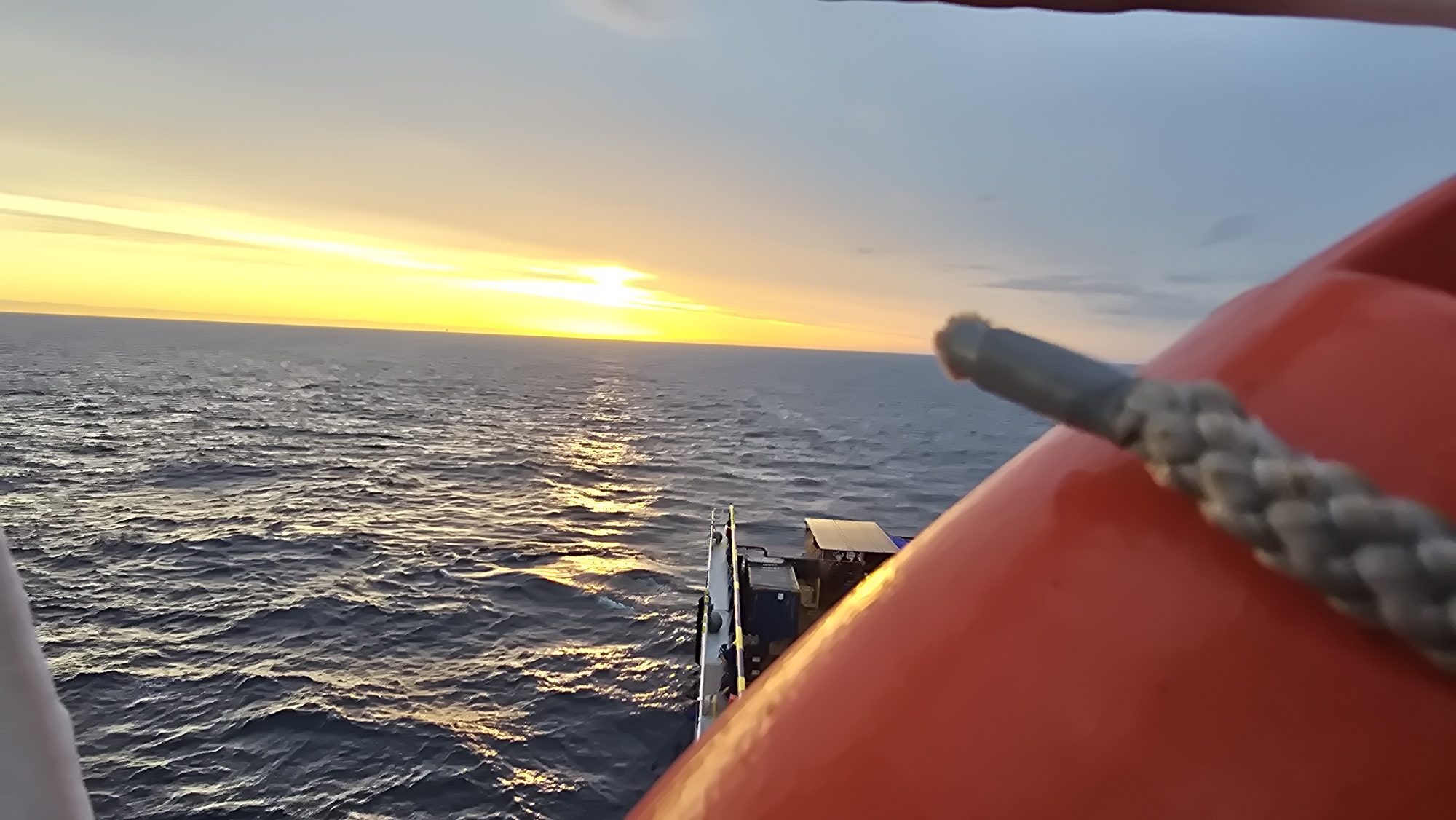 The image shows a view of the ocean from a boat, with a sunset on the horizon. A red safety device with a gray rope attached is in the foreground. The boat itself is visible towards the center of the frame. The location is Unknown, Unknown.