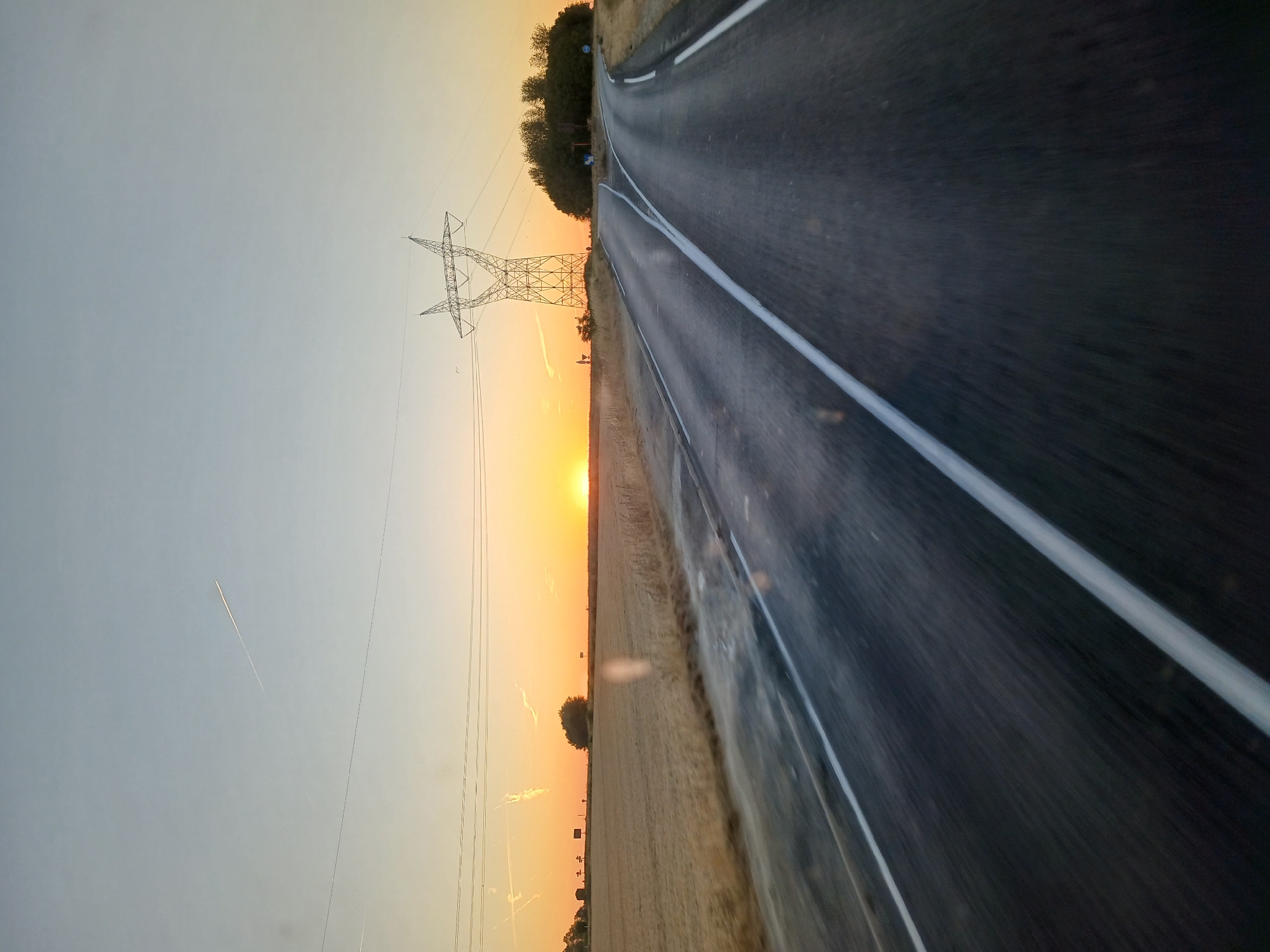 An asphalt road curves to the right, bordered by a cultivated field on the left and a stand of trees on the right. A power line tower stands prominently on the right side of the road, with power lines stretching across the sky. The sky is a gradient of pale blue to orange, with the sun low on the horizon, indicating either sunrise or sunset. A contrail from an airplane is visible in the upper left quadrant of the sky. The image is captured from a moving vehicle, evidenced by the motion blur on the road and the edges of the frame. The location is identified as Penchard, France.