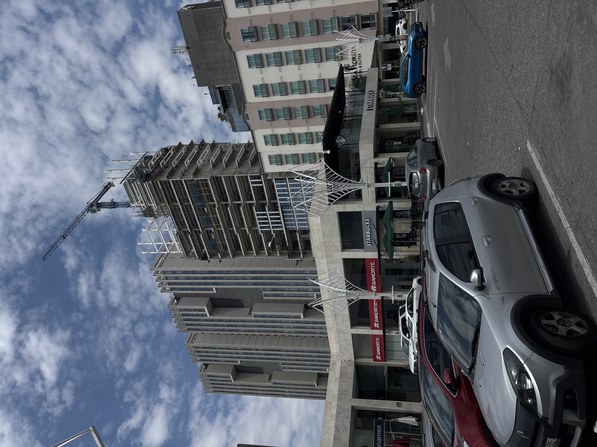 The image captures a bustling urban scene in San Bernardino Tlaxcalancingo, Mexico. A modern high-rise building under construction dominates the view, with a crane extending toward the sky filled with scattered clouds. Adjacent to the construction site, a multi-story building houses a Starbucks, identifiable by its logo, and a Banorte bank branch. Several cars are parked along the street, including a silver Renault Stepway and a blue vehicle. The overall impression is of a vibrant, developing area within the city.