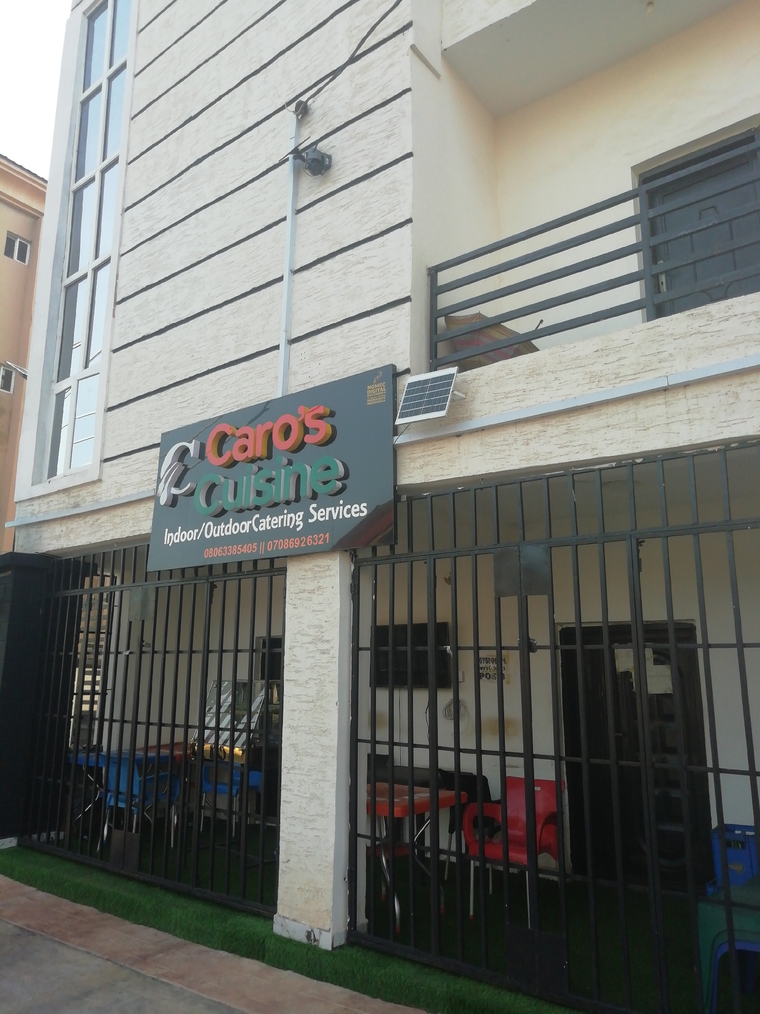 The image shows the exterior of Caro's Cuisine, an indoor/outdoor catering service located in Enugu, Nigeria. The building is a two-story structure with a textured, light-colored facade and horizontal black stripes. A large sign displays the restaurant's name and contact information. A solar panel is mounted above the sign. Security cameras are visible on the wall. A black metal fence surrounds an outdoor seating area with tables and chairs. Artificial turf is visible near the base of the fence. A partial view of a neighboring building is seen on the left.