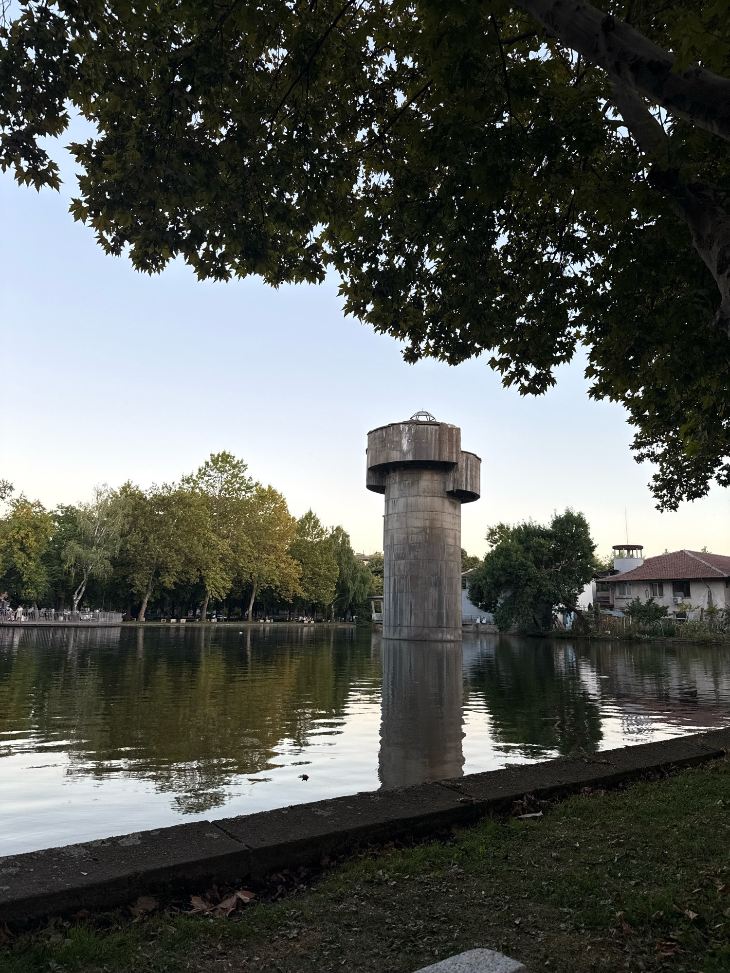 A water tower stands in the middle of a lake surrounded by trees and parkland in Montana, Bulgaria. The tower is made of concrete and has a cylindrical base with two larger concrete structures at the top. The lake's surface is calm, reflecting the trees and the sky. In the background, there are more trees, some buildings, and a clear sky. The foreground shows grass and a concrete walkway.