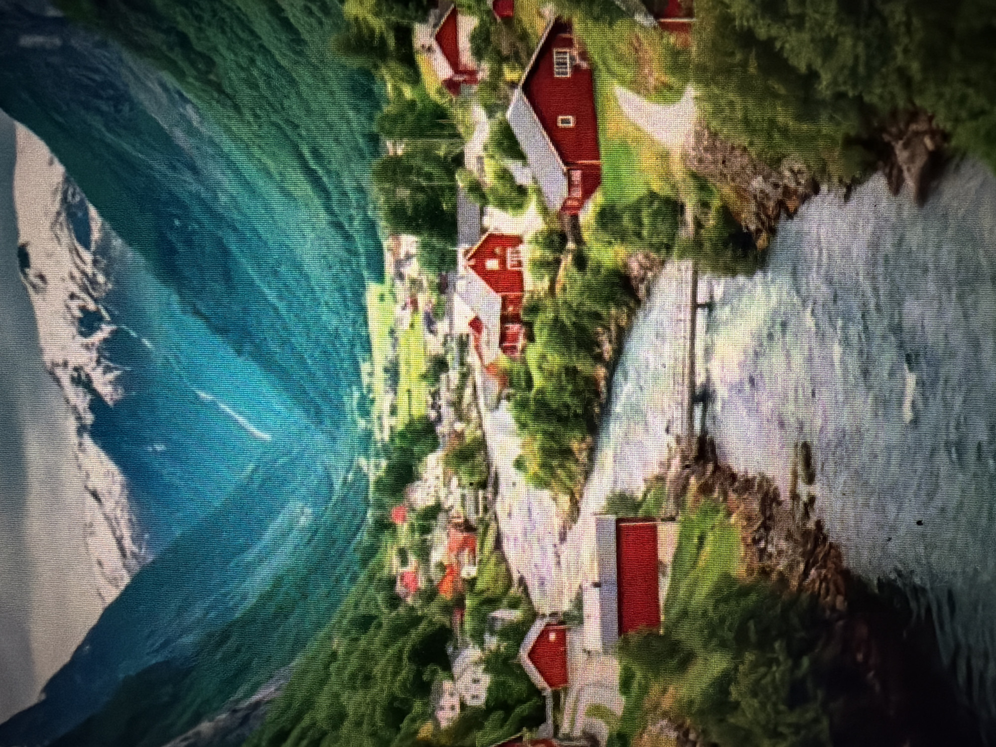 The image depicts a picturesque Norwegian fjord landscape. Towering, snow-capped mountains form the backdrop, with steep, verdant slopes descending into a valley. A village of traditional red-roofed houses is nestled along the banks of a winding, clear river that flows through the valley. The river is crossed by a small bridge. The scene is bathed in natural light, highlighting the lush greenery and the textures of the mountains and water. The image does not show the city of Baan Mae Nam, Thailand, as it depicts a Norwegian landscape.