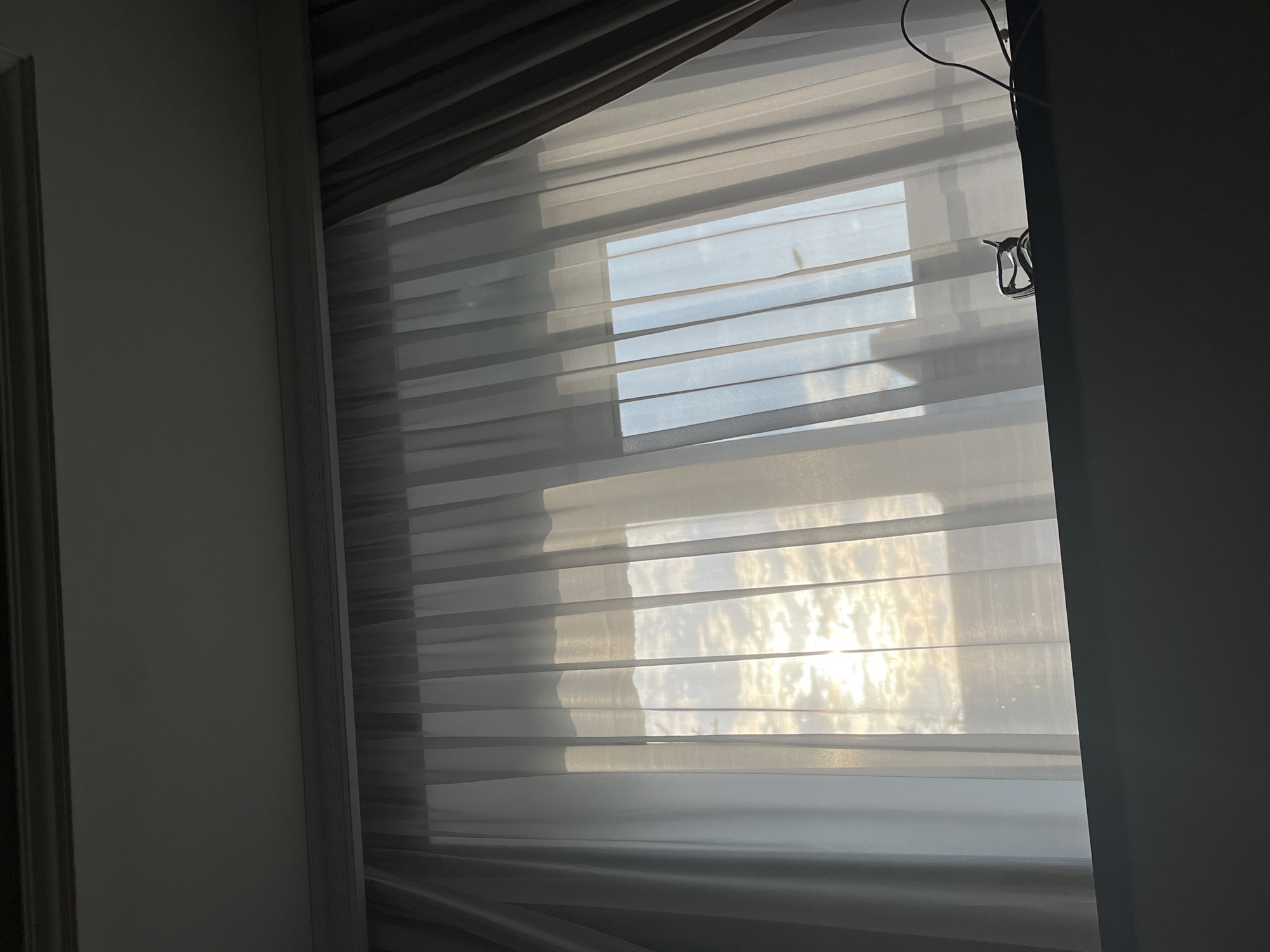 The image shows a window with sheer white curtains pulled partially closed, revealing a sky with scattered clouds illuminated by sunlight. A pair of eyeglasses and a charging cable are visible on the windowsill. The scene appears to be indoors, with a dark ledge in the foreground and the ceiling with ornate molding at the top. The lighting suggests it might be morning or late afternoon. There are no people or specific identifiable landmarks of Astrakhan, Russia visible in the image.