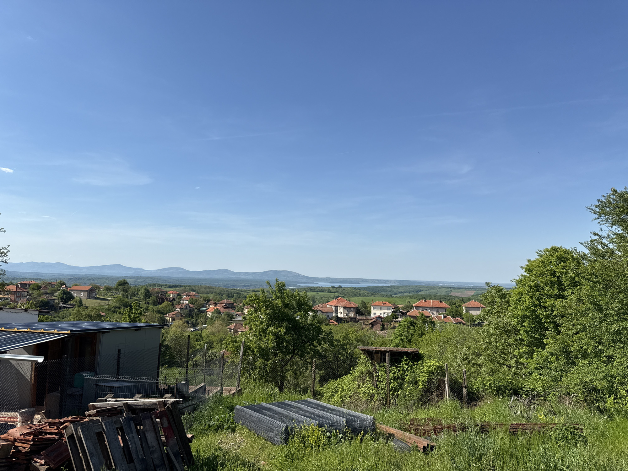 The image depicts a landscape in Borovtsi, Bulgaria. In the foreground is a cluttered area with building materials, including a stack of metal mesh, wooden planks, and red roof tiles. A metal shed is also visible. The mid-ground features a residential area with houses with red tile roofs interspersed with trees and green vegetation. In the background, a range of hills and a body of water are visible beneath a clear blue sky with thin clouds. The scene is taken during daylight hours.