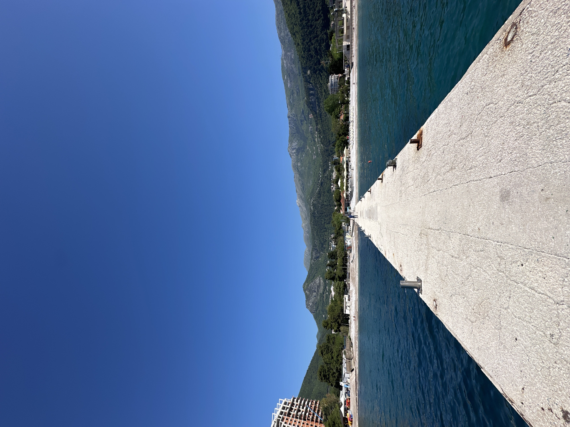 A concrete pier extends from the shore into the sea on a clear, sunny day. The sky is a bright, unblemished blue. The pier, wide at its base and appearing to taper off into the distance, has metal fixtures along its edges.
On the left of the pier, the coastline stretches along a bay with trees, buildings, and mountains rising sharply in the background. A beachfront with various structures and trees lies between the water's edge and the lower slopes of the mountains. The water is a deep blue. In the foreground near the lower-left corner, a taller structure, possibly a multi-story building or hotel under construction, is visible.
The image appears to be taken in or near Budva, Montenegro, given the coastal setting, the type of architecture, and the mountainous terrain. The presence of a pier alongside a beach indicates that the location is likely a coastal recreational area within Budva.