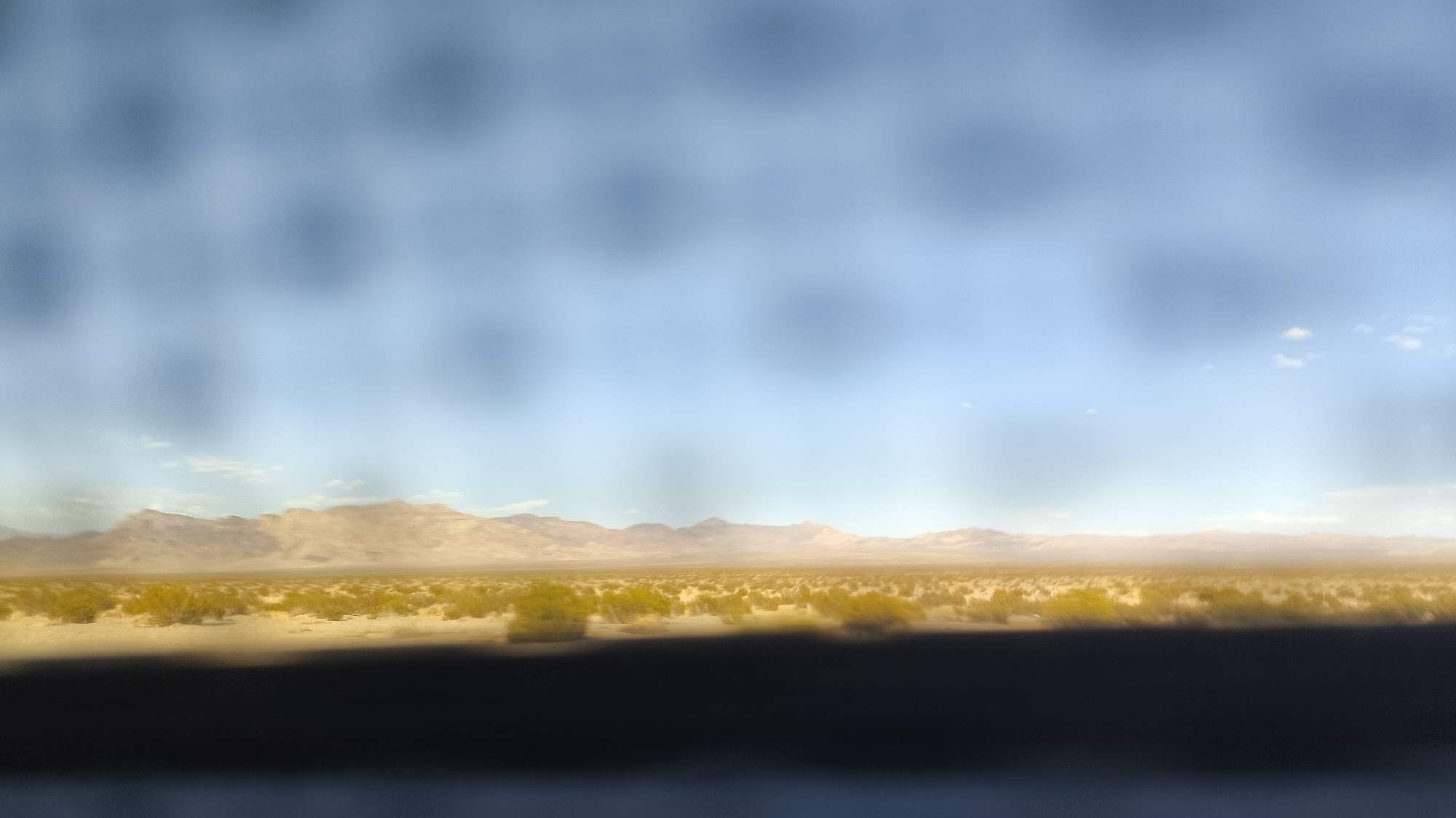 The image depicts a vast, arid landscape under a pale blue sky with scattered clouds. The foreground is dominated by a dark, blurred strip, likely representing a road or vehicle interior. Beyond this, a wide expanse of dry, light-colored ground is dotted with sparse, green desert vegetation. In the distance, a range of beige mountains stretches across the horizon, their peaks softened by atmospheric haze. The overall impression is one of a desolate, sun-drenched desert environment. The image does not contain any people, actions, or interactions. The location cannot be identified as Renoville, United States, as there is no visual evidence to support this claim.