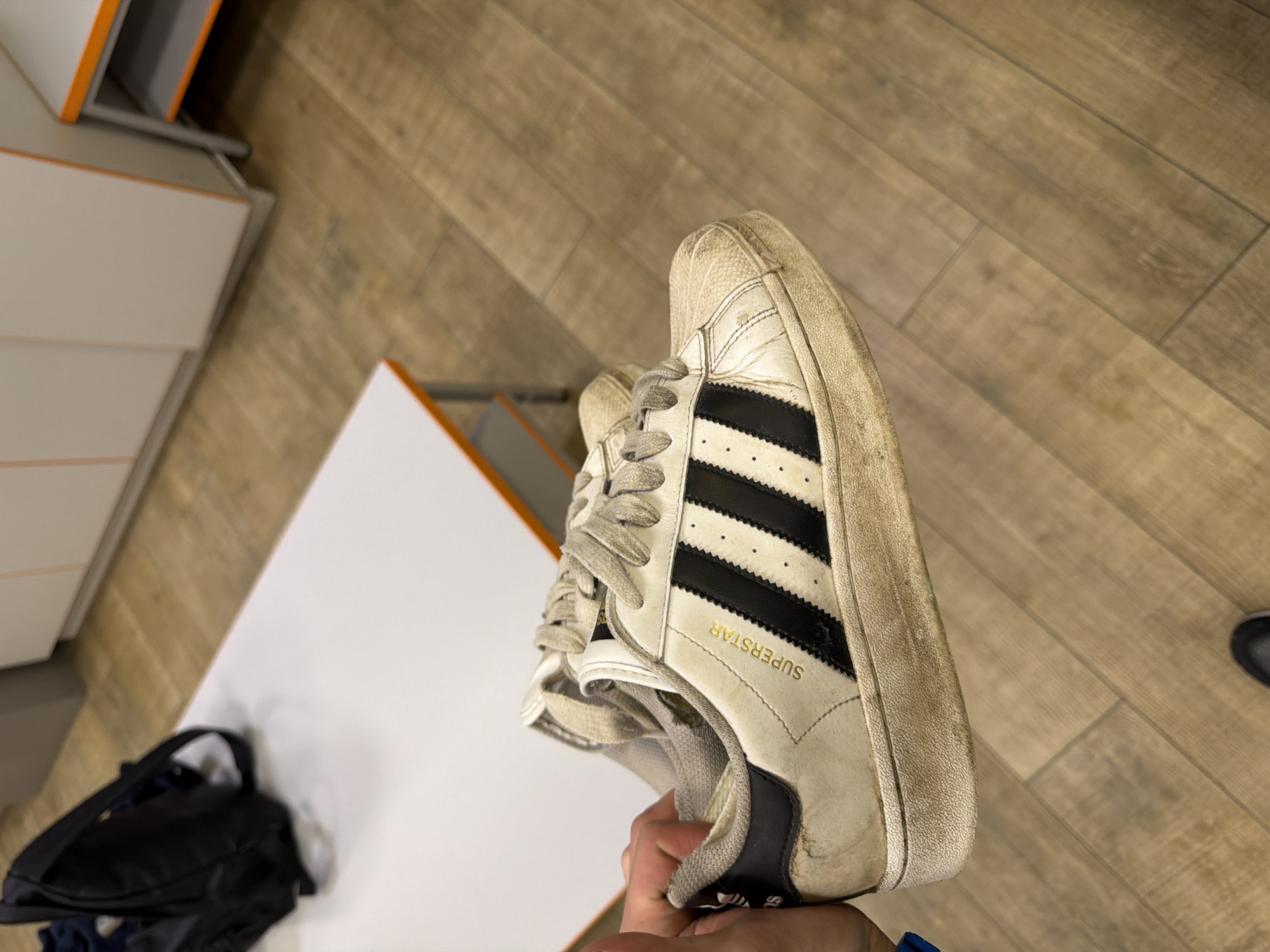 The image shows a pair of dirty white Adidas Superstar sneakers being held by a person's hand. The sneakers have black stripes and the word "SUPERSTAR" is written in gold on the side. The background consists of a wooden floor and what appears to be a white table and some cabinets, suggesting an indoor setting. There is no indication of the location being Nizhny Novgorod, Russia.