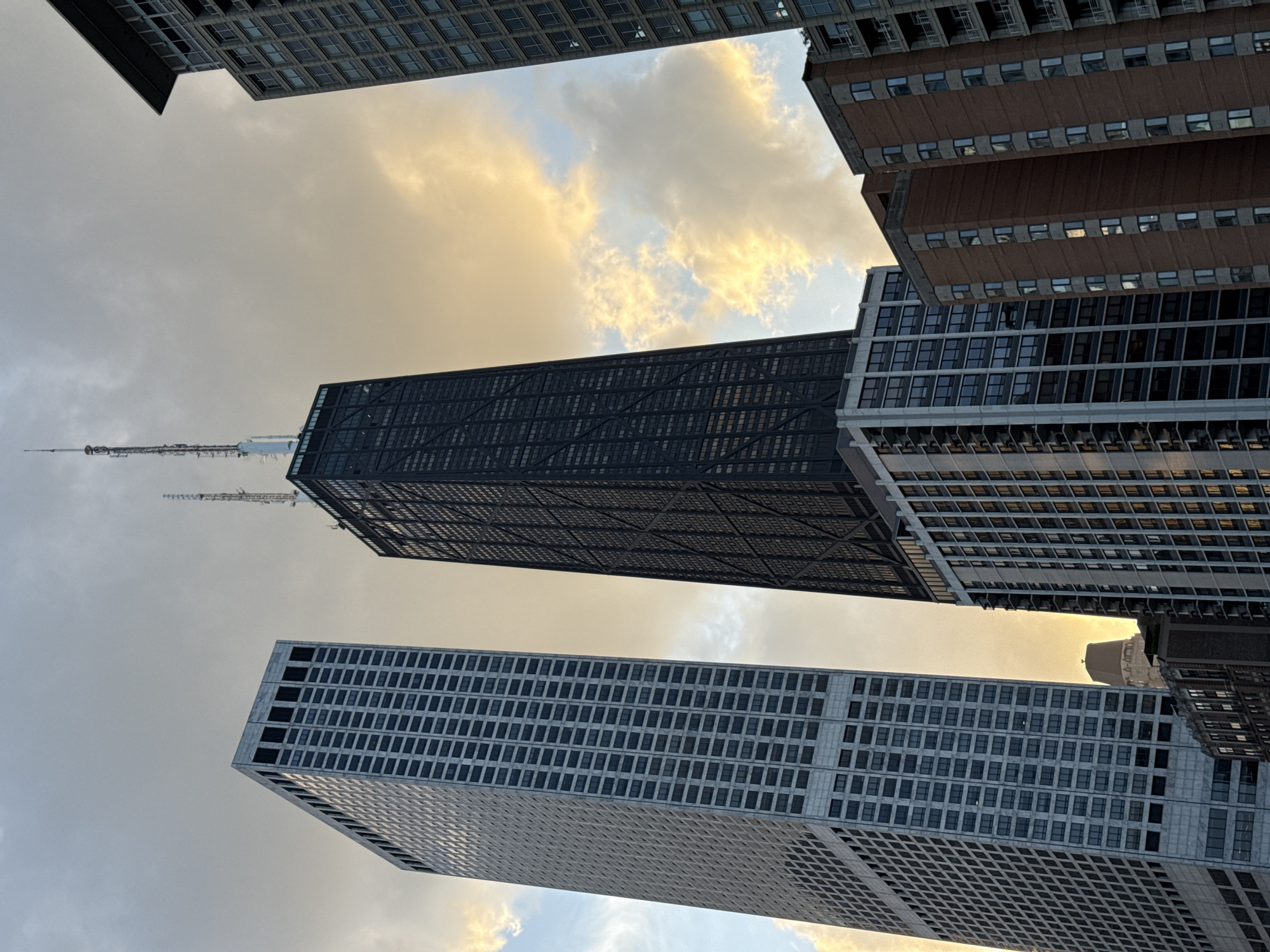 This image captures a low-angle view of several skyscrapers in Chicago, Illinois, USA, against a dramatic cloudy sky illuminated by golden sunlight. The most prominent structure is the John Hancock Center, a dark, X-braced skyscraper with a distinctive antenna at its peak, located in the heart of the city. To its left stands a tall, rectangular, light-colored skyscraper with a grid-like facade. To the right and in the foreground, a mid-rise building with a brick facade and numerous windows is visible. Further to the right, another modern high-rise building with a glass facade completes the urban canyon effect. The sky is a mix of gray clouds with patches of warm, golden light, suggesting either sunrise or sunset. No people or vehicles are discernible in the frame.