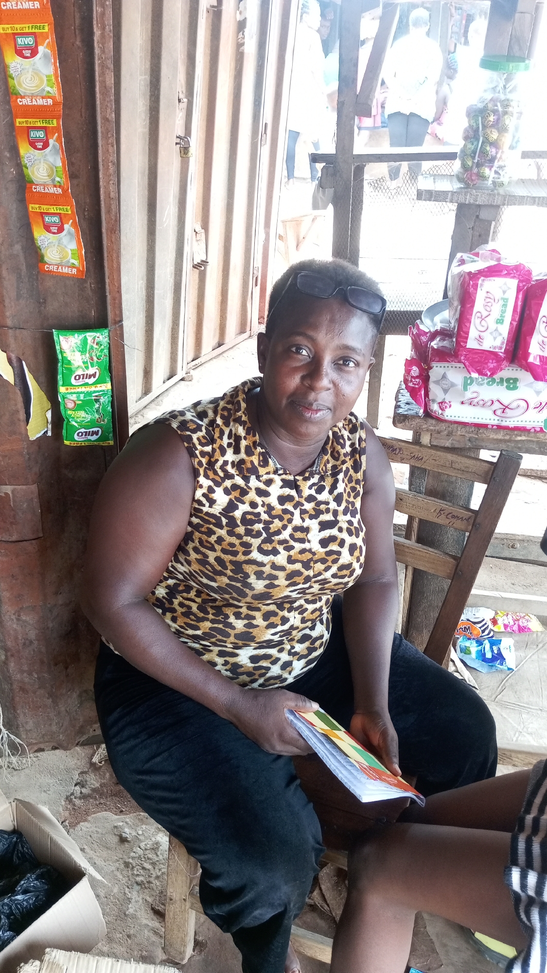 The image shows a woman seated on a wooden chair outside what appears to be a small shop or stall in Nkpor, Nigeria. She is wearing a leopard-print top and black pants, with sunglasses perched on her head, and is holding a booklet or stack of papers in her hands. Behind her, on a wooden wall, are packets of Kivo creamer and Milo drink mix displayed for sale. To her right, on a separate wooden structure, there are packets of bread with the brand "de Rosy" visible, and a jar filled with candies. The background suggests an outdoor setting, possibly a market or street scene. A cardboard box sits on the ground near the woman.