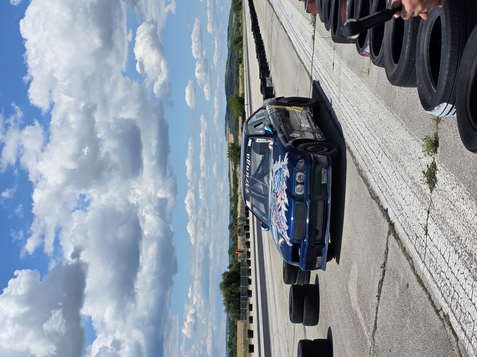 A dark blue drift car with a colorful dragon decal on the hood is parked on a paved race track in Akatsievo, Bulgaria. The car has a roll cage and racing tires. In the foreground, stacks of used tires are piled on the left and right sides of the track. A person's hand is visible on the right, holding a wrench. The background features a flat, open landscape with trees and hills under a bright blue sky with scattered white clouds. A digital timer displaying "00.000" is visible in the distance.