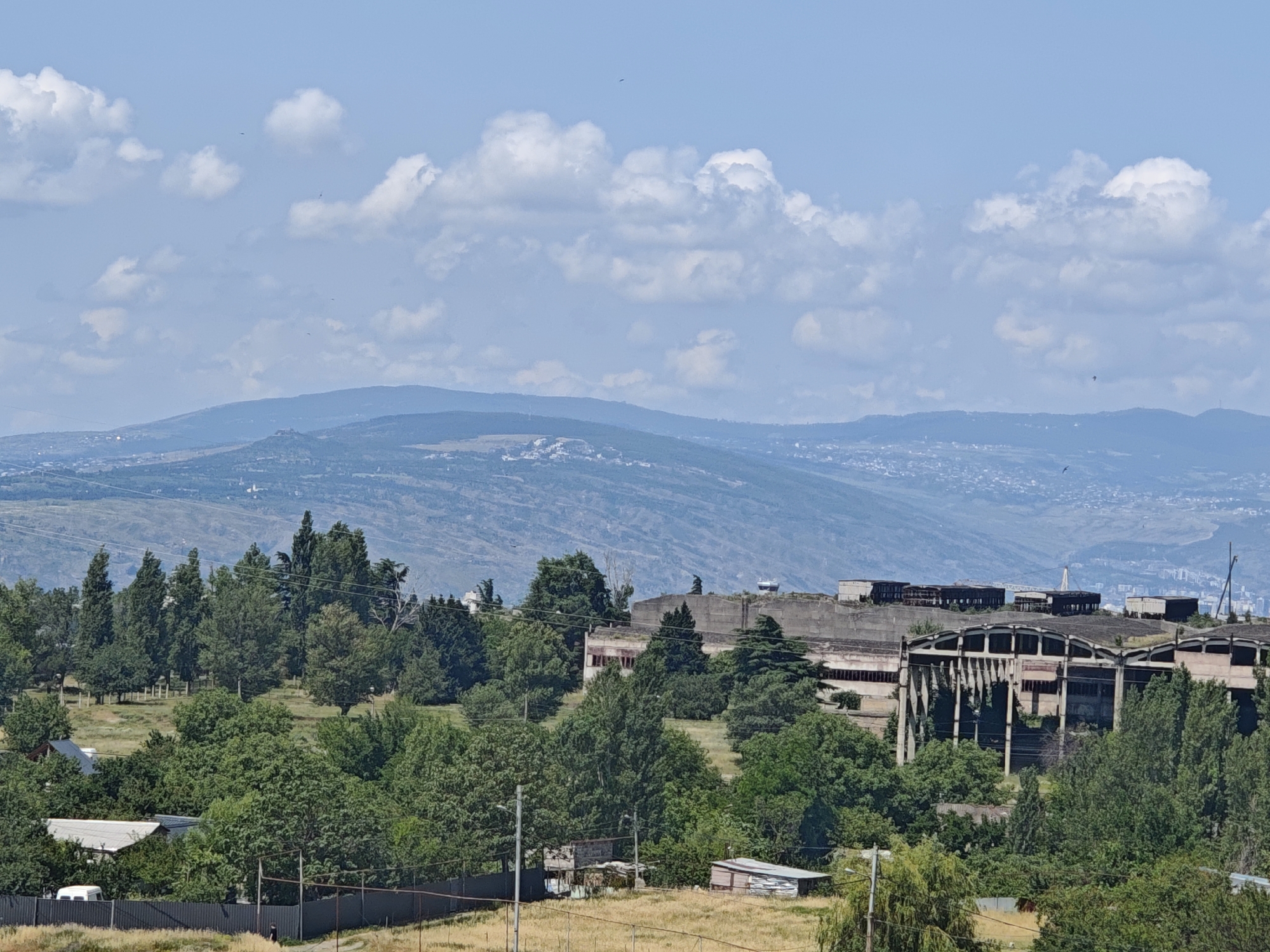 This is a high-angle, wide shot of an abandoned industrial complex on a hillside in Tbilisi, Georgia. The foreground shows a chain-link fence with a small white van parked behind it. A dirt path leads away from the fence towards some small sheds in the middle ground. The middle ground also features a variety of lush green trees, some taller and more mature, others smaller and shrub-like.
Dominating the mid-right of the frame is the derelict industrial structure. It appears to be a large, multi-level building with a concrete facade and a distinctive arched roofline that has partially collapsed in places. Many of the windows are broken or missing. The building is surrounded by trees, with some growing directly through the structure itself.
The background reveals rolling hills covered in a mixture of trees and scattered buildings, indicating the urban sprawl of Tbilisi. Further in the distance, larger, hazy blue mountains form the horizon. The sky above is a bright, clear blue with scattered white cumulus clouds, and a few small dark specks, possibly birds, are visible in the sky. The overall impression is one of decay and nature reclaiming an industrial site.