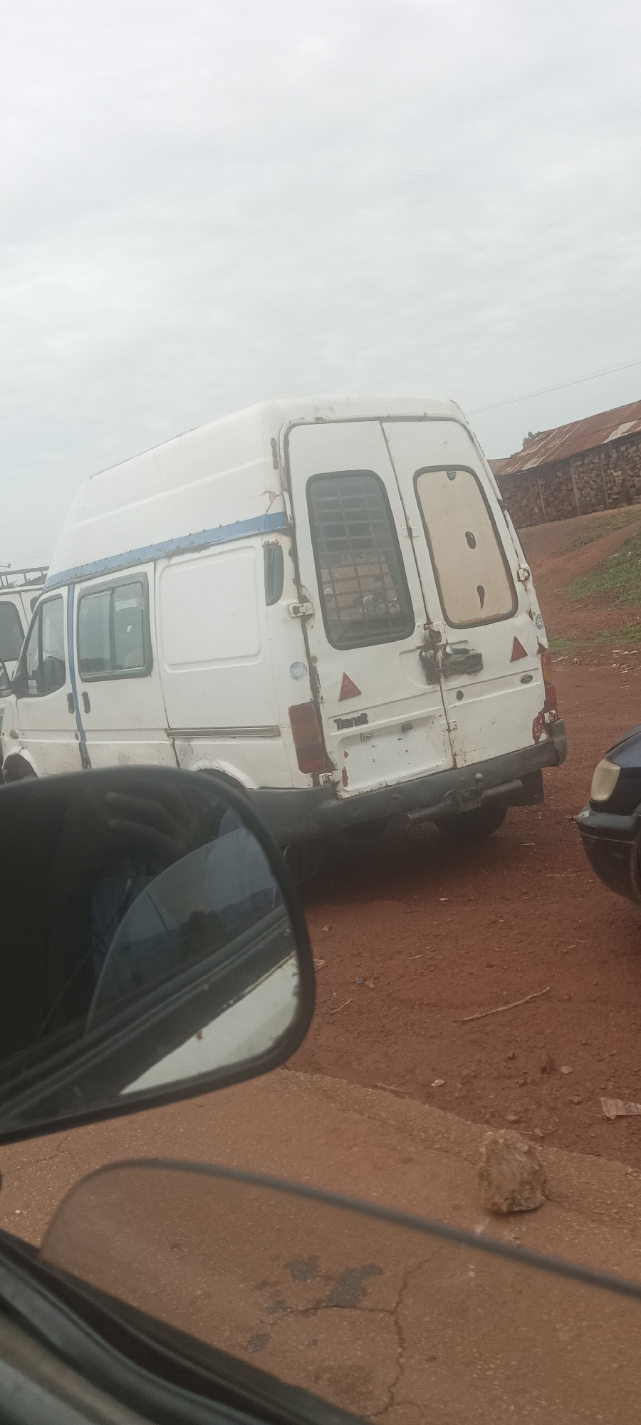 A white Ford Transit van is parked on a red dirt road. The van appears to be old, with visible damage and rust. The rear doors are different - one has a window with metal bars, the other has an empty window frame. The word "Transit" is visible on the back. The van is partially obscured by the side mirror of another vehicle. In the background, there is a building with a rusty corrugated iron roof. The location is rural or semi-rural, and it is likely in Durumi, Nigeria, as the context suggests.