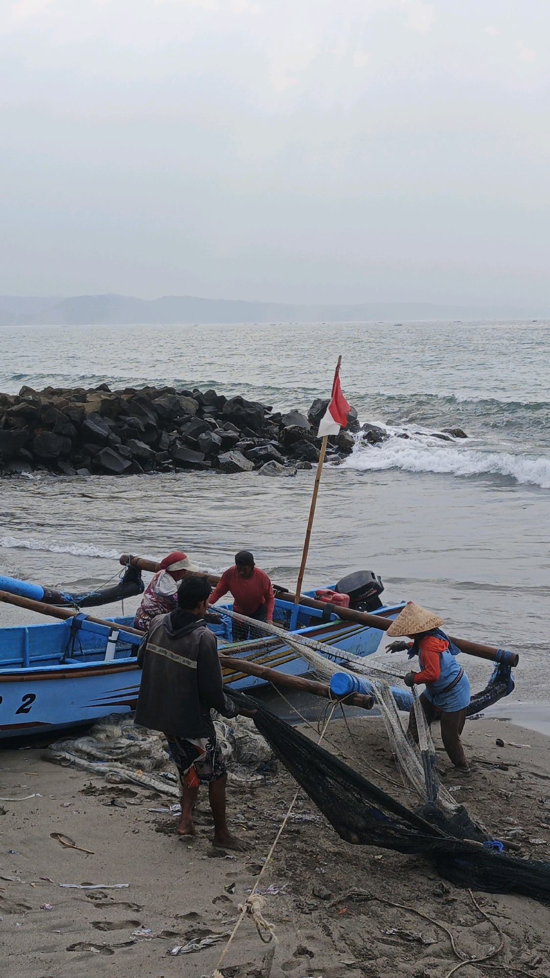 In Desa Pagandaran, Indonesia, on a sandy beach near the ocean, a group of fishermen are preparing their fishing nets. A blue boat is pulled ashore, with a red and white Indonesian flag flying from a wooden pole attached to it. Four men are visible. One man, wearing a dark hooded jacket and patterned shorts, stands on the beach near the stern of the boat, holding a black fishing net. Another man, wearing a red shirt, is inside the boat. A third man, wearing a patterned headscarf and what appears to be a vest, is also in the boat. The fourth man, wearing a conical straw hat and an orange shirt over a blue garment, is on the beach near the bow of the boat, working with a fishing net. The sea is visible in the background, with choppy waves and a rocky breakwater. The sky is overcast. The sand is littered with debris, including plastic.