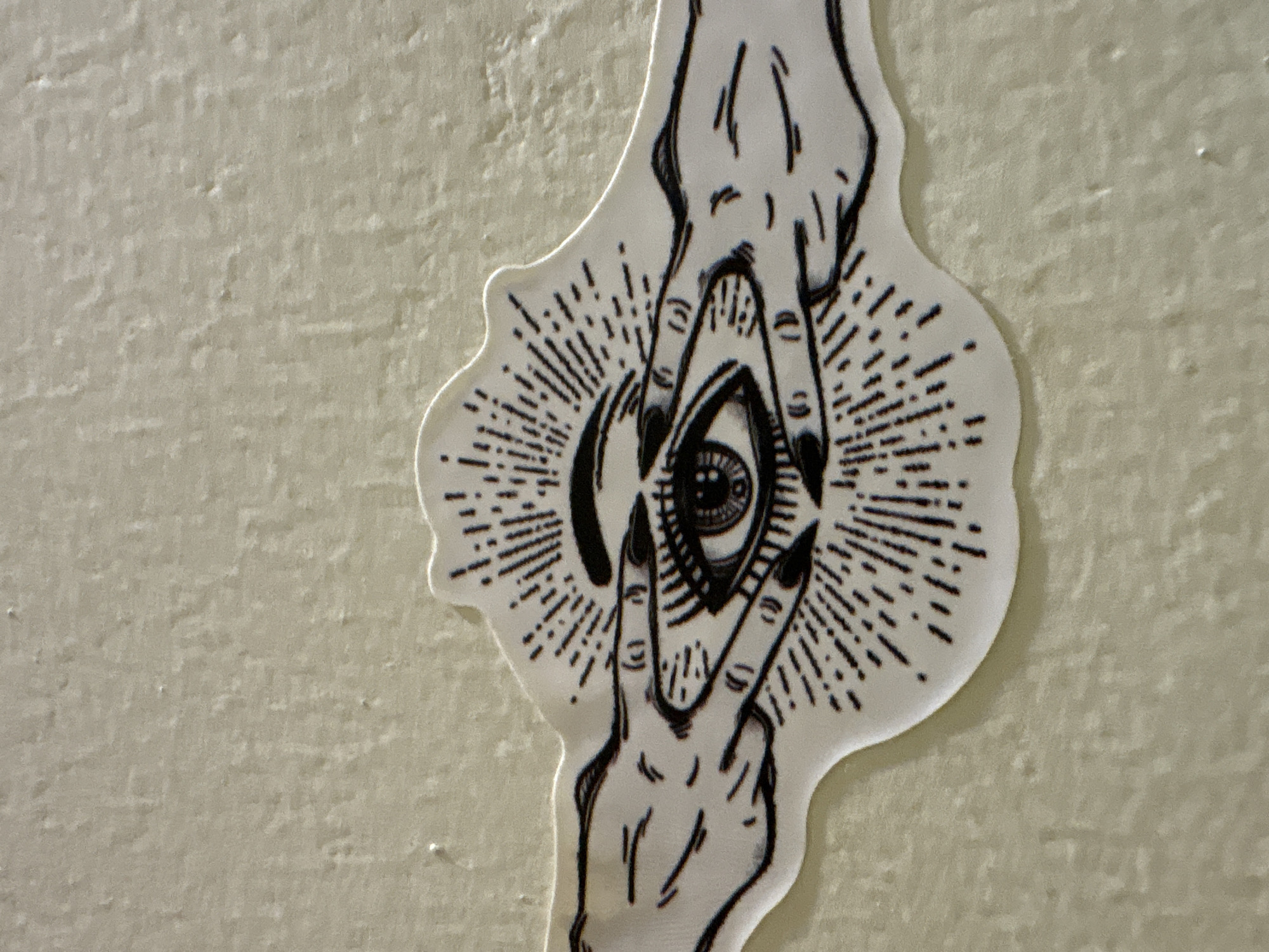 A close-up photograph captures a sticker with an eye design adhered to a light-colored, textured wall. The sticker features a stylized eye with eyelashes and a pupil, surrounded by radiating lines, suggesting a sun or a mystical symbol. Two hands, drawn in black ink, frame the eye from the sides, with fingers extended and pointing towards the center. The sticker has a white border and a die-cut edge. The wall appears to be an interior surface, possibly a home or a commercial establishment, in Ashvem, India.