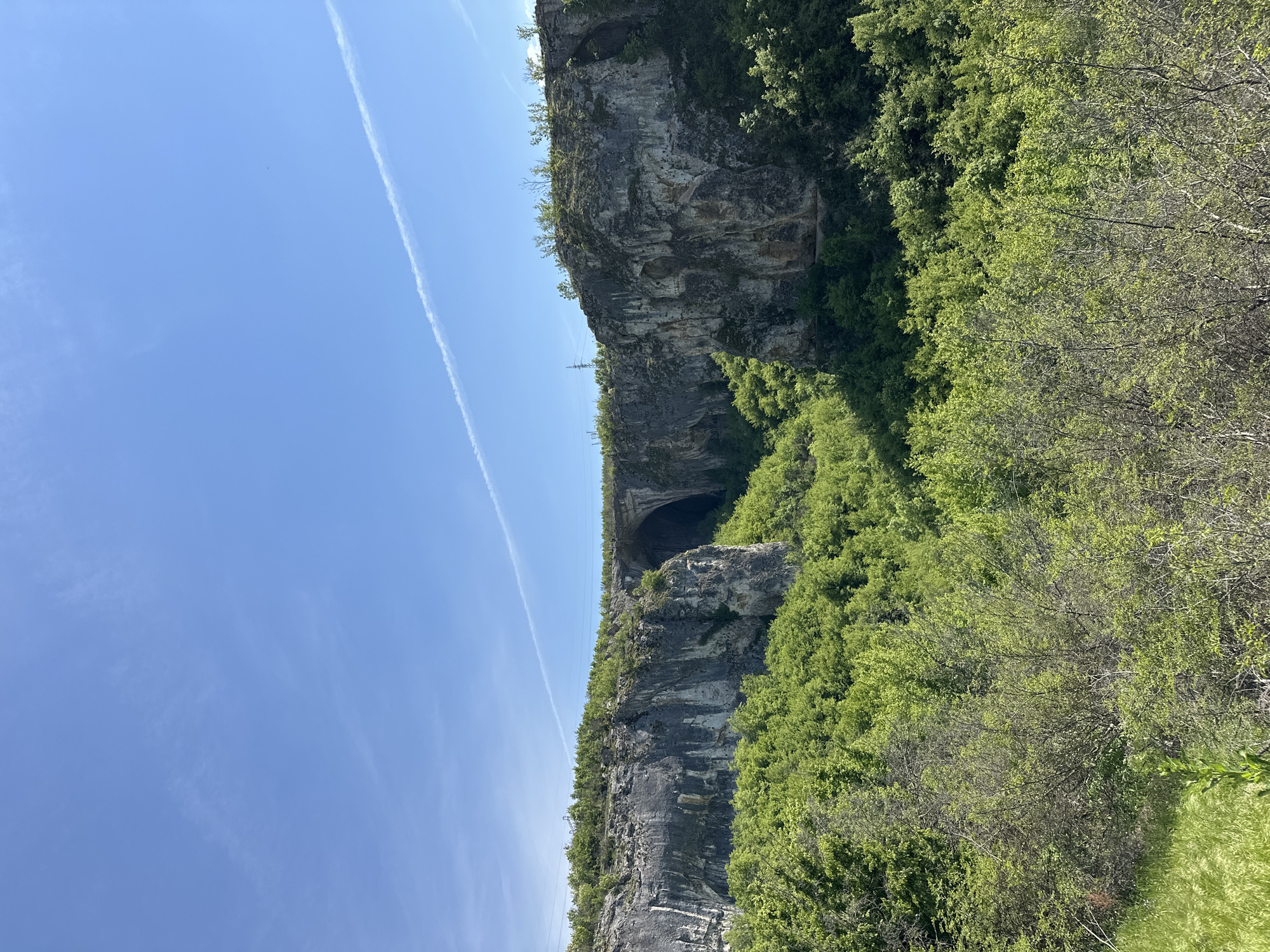 The image shows a cliff face in Karlukovo, Bulgaria. The cliff is composed of gray rock and covered in green vegetation. There is a large cave opening in the cliff face. The sky is blue with a contrail.