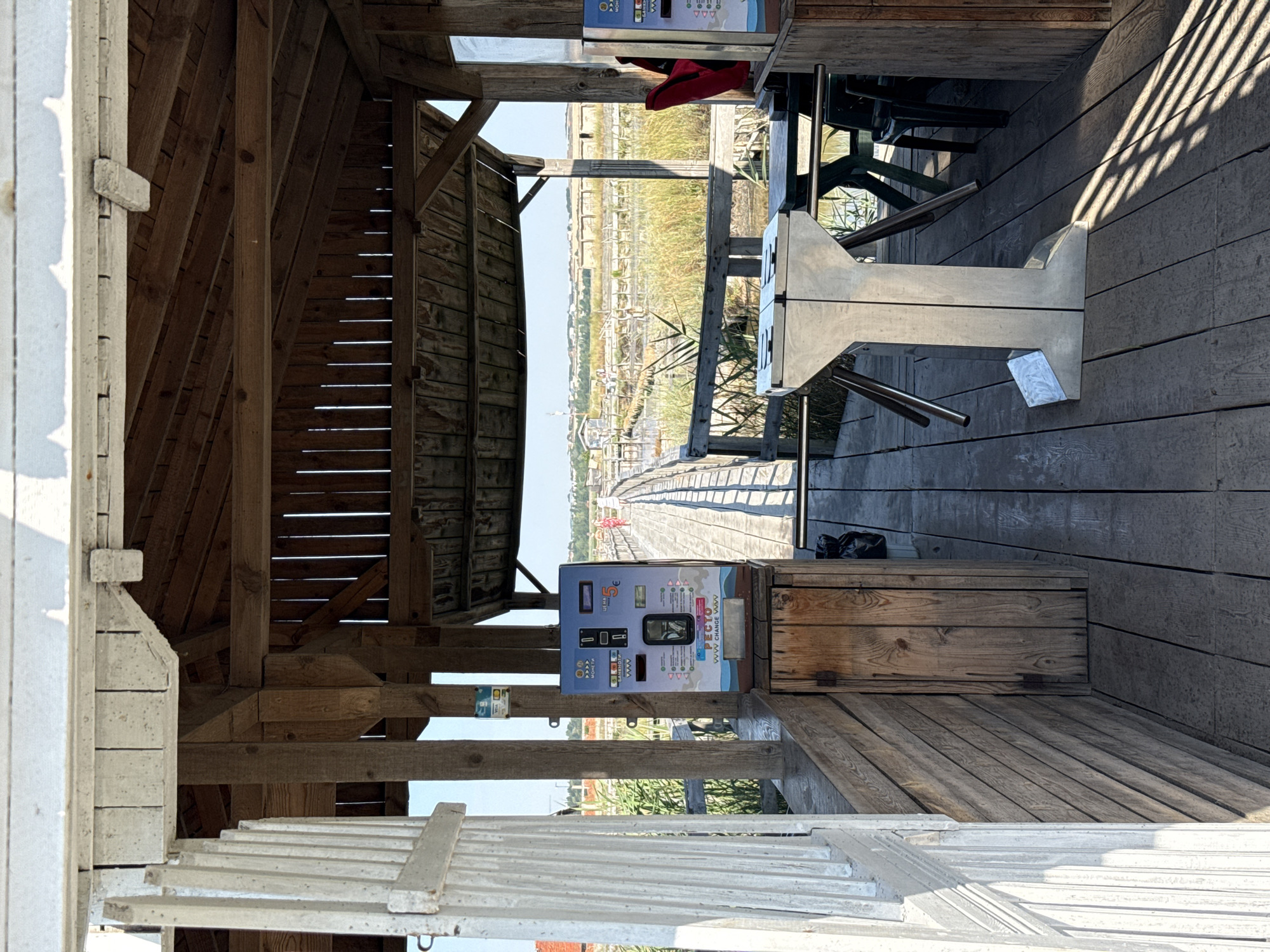 The image shows an outdoor wooden structure, likely an entrance or ticket booth, at the Atanasovsko Lake salt pans in Burgas, Bulgaria. In the foreground, a white, weathered wooden gate is partially open on the left. The structure has a wooden roof and floor, supported by wooden posts. A blue ticket vending machine with a display screen and slots for money is prominently placed on a wooden counter to the left of the entrance. Below the machine is a small wooden cabinet with a hinged door.
In the middle ground, a silver turnstile stands on the wooden floor, serving as an entry point. Behind the turnstile, a wooden walkway or bridge extends into the distance, leading over a body of water with reeds and marshy vegetation. The landscape appears to be a salt pan with structures visible in the background. A person in a red top is partially visible to the right of the vending machine, near a table and chairs. The lighting suggests it is daytime, with sunlight casting shadows.