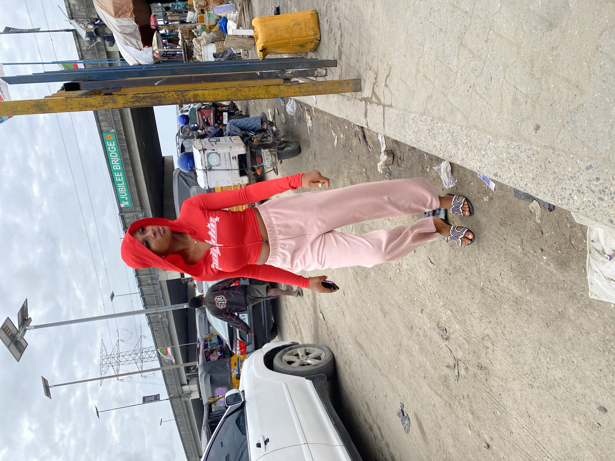 A young woman stands on a city street in Lekki, Nigeria. She wears a red hooded crop top and light pink sweatpants. Her feet are adorned with sandals, and she holds a phone in her hand. The environment includes a bridge labeled "JUBILEE BRIDGE," cars, motorcycles, and street clutter. The overall impression is a snapshot of everyday life in a busy urban setting.
