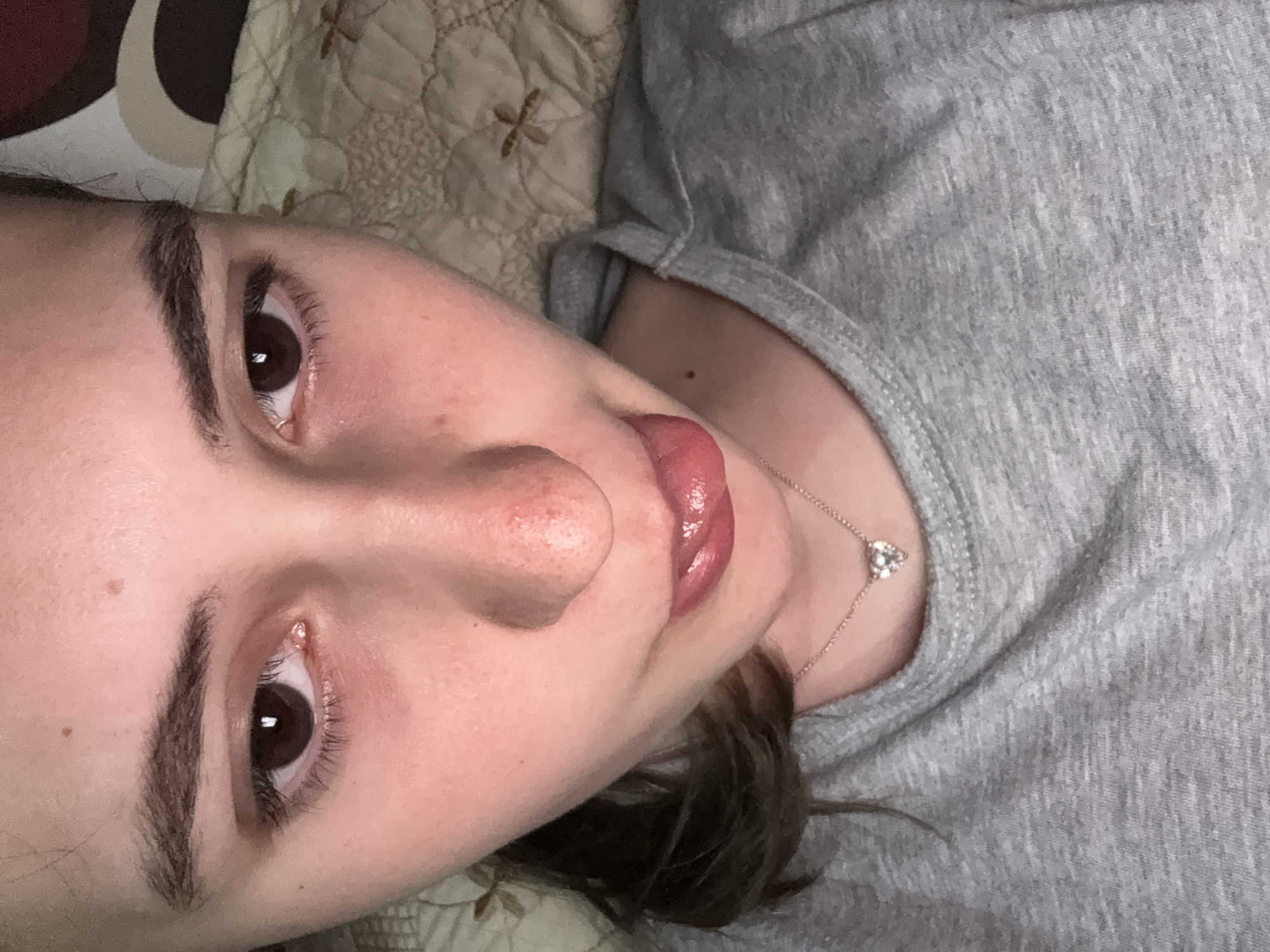 A young, fair-skinned person with dark brown eyes is lying on a quilted, beige-toned surface, possibly a bed. The person is wearing a gray T-shirt and a delicate necklace with a small, triangular pendant. The person's lips are slightly parted, and their tongue is visible. The image is a close-up, focusing on the person's face and upper body. There is no indication of a location in Panagyurishte, Bulgaria, or any other specific location.