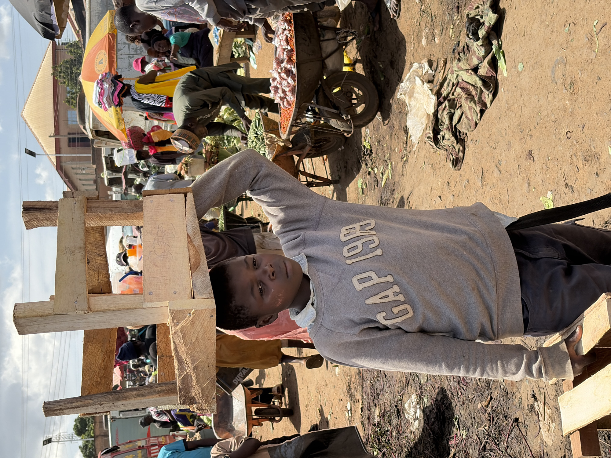 This image captures a vibrant outdoor market scene in Gura Top, Nigeria, bathed in bright daylight. The ground throughout the market appears to be unpaved dirt, showing signs of wear and activity.
The primary subject in the foreground is a young person, likely a boy, standing next to a rudimentary wooden structure, possibly a stall frame or barrier. He looks directly at the camera with a neutral expression. He is dressed in a grey hooded sweatshirt with the text "GAP 1969" prominently displayed in white letters across the chest, and dark-colored trousers. His right arm rests on the wooden structure.
The midground reveals the bustling market environment. Numerous individuals are seen moving about or congregating around various market setups. A bright yellow and red market umbrella provides shade for one of the stalls. Adjacent to this, a vendor's cart, fitted with a bicycle wheel, holds a large quantity of what appear to be small, red dried goods, possibly peppers. Other people are visible engaged in market activities, such as browsing or selling; one individual wears a bright yellow top, and another a pink cap. Various merchandise, including textiles and produce, are spread out. The word "WANG" is faintly visible on a bag in the lower left portion of the image.
In the background, several buildings with light-colored roofs are visible, along with a utility pole and overhead wires stretching across a clear, bright sky. The overall scene conveys the lively atmosphere of a typical informal market.