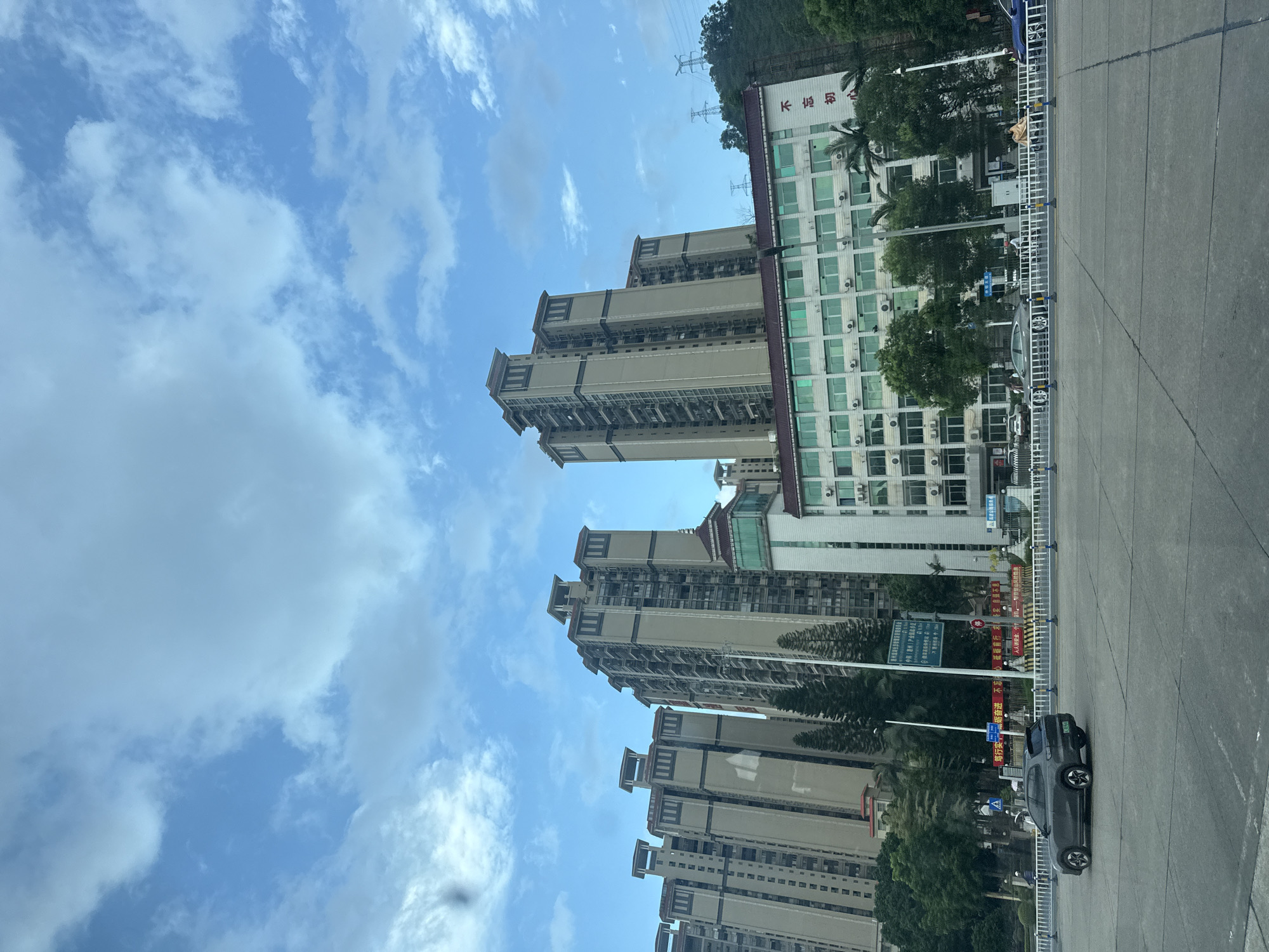 The image depicts a street scene in Huizhou, China, with multiple tall residential buildings dominating the background. These buildings are characterized by their light-colored facades, numerous windows, and distinct tiered rooftops. In the middle ground, a lower, multi-story building with green-tinted windows is visible, bearing Chinese characters on its side. A dark-colored SUV is driving on the road in the foreground, with other vehicles and pedestrians present further down the street. The sky is blue with scattered white clouds. The overall environment appears to be a well-developed urban area.