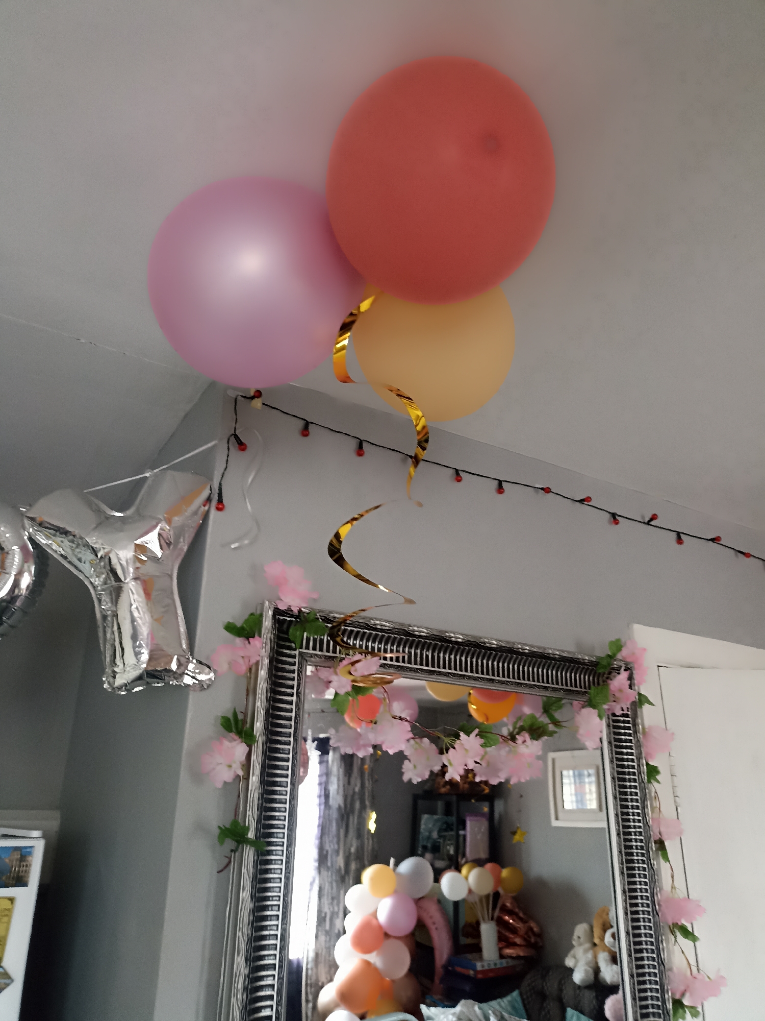 This is an indoor scene, likely a living room, with a celebration theme. The focal point is a large, ornate mirror with a silver frame, decorated with a string of pink artificial cherry blossoms. Suspended from the ceiling are three large, round balloons: one pink, one coral, and one orange. A silver foil balloon, shaped like the letter "Y", is also visible, attached to the wall. There are also strings of small, red-lighted fairy lights running along the wall above the mirror. The mirror reflects a collection of smaller balloons, some in a bouquet and others scattered, along with some stuffed animals. The walls are painted a neutral grey. The location is identified as Annagry, Ireland, but no specific details about the city are visible in the image itself. There are no people present in the frame.