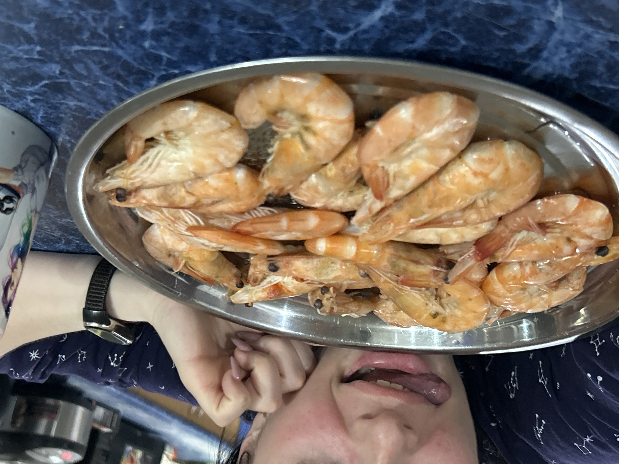 A photograph shows a silver oblong platter filled with cooked shrimp. The platter is oriented horizontally in the frame. Below the platter, a person's face is visible, with the tongue extended. A wrist with a black watch is visible to the left of the platter. The person is wearing a dark blue shirt with white constellation patterns. The background appears to be a dark blue surface, possibly a table. A partially visible white mug with decorations sits to the left of the platter. The location of Rusalevka, Russia, cannot be determined from the image.