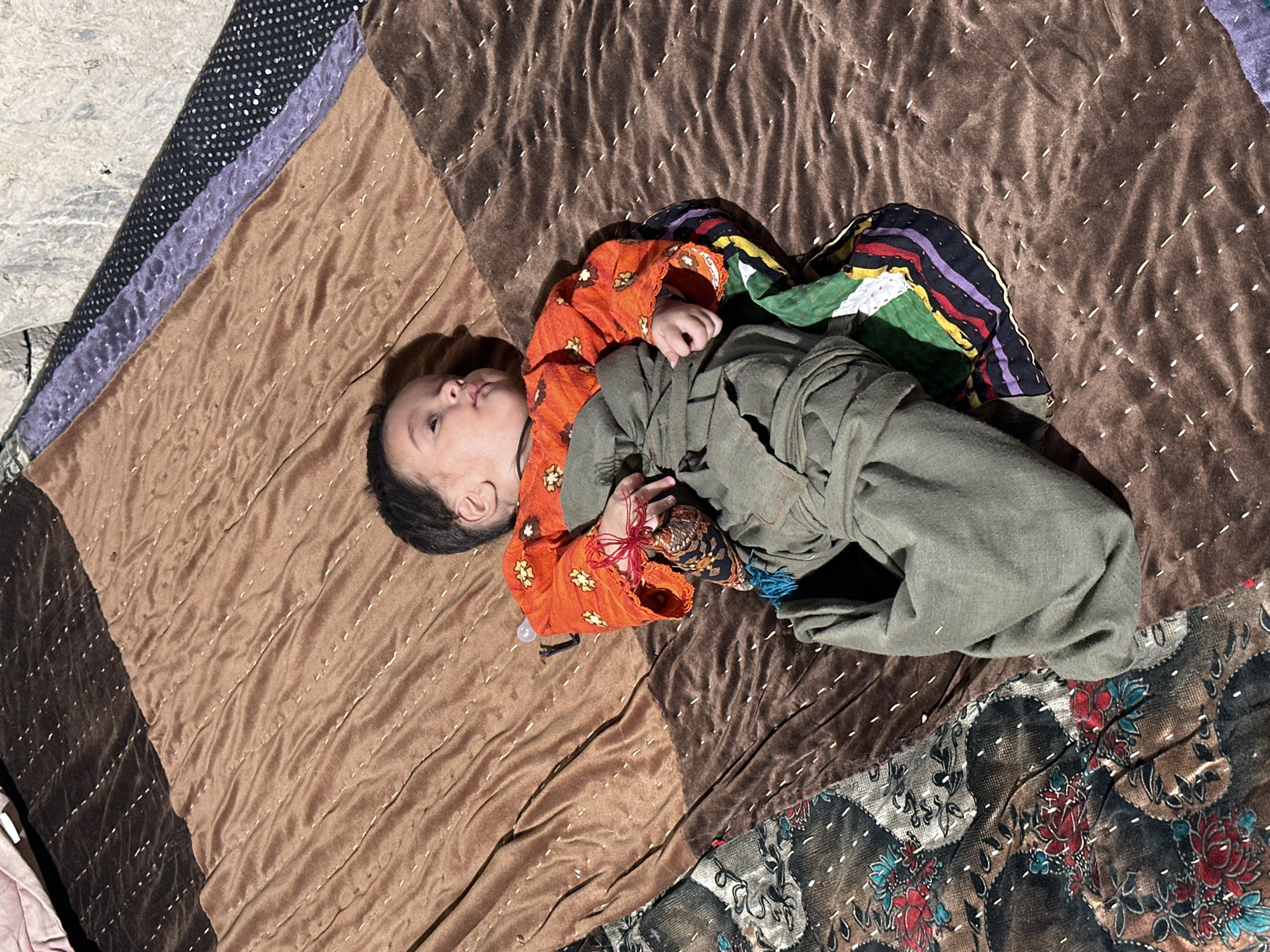 A baby lies on a patterned quilt outdoors in Quetta City Tehsil, Pakistan. The baby is wrapped in loose, olive-green clothing over an orange shirt with floral patterns. The baby’s head is turned to the left. The quilt underneath features varying patterns including brown and tan panels with visible stitching, a black and silver polka dot section, and a floral-patterned corner. Some of the quilt lies on a concrete surface. The lighting suggests direct sunlight.