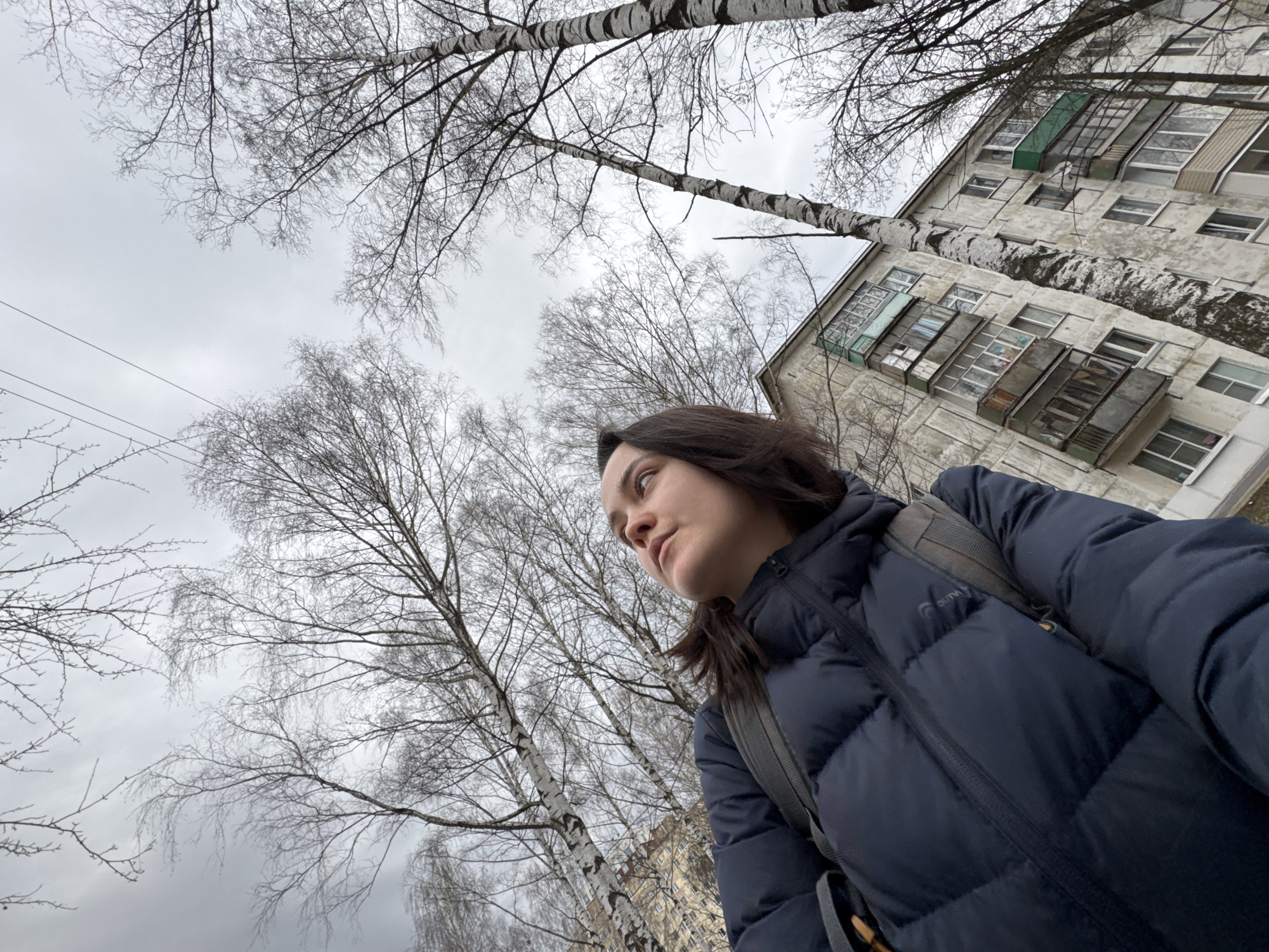 The image presents a low-angle, upward-looking perspective of a woman outdoors in an urban or suburban setting, likely Izhevsk, Russia. The woman, with dark hair, is seen from the chest up, looking intently upwards and to her right. She wears a dark blue, padded puffer jacket, on which a brand logo, appearing to be "OUTVENTURE," is visible on her left chest. A grey backpack is slung over her shoulders. Her expression is neutral and observant.
The sky is heavily overcast, casting a soft, muted light over the scene. The weather appears cool or cold, indicated by her warm attire and the bare branches of the numerous deciduous trees, suggesting late autumn, winter, or early spring. Tall, slender birch trees with characteristic white and dark bark patterns dominate the composition, their intricate network of leafless branches filling a significant portion of the frame against the grey sky.
To the upper right, a multi-story residential building with a weathered, light-colored facade is partially visible, its architectural style, including multiple windows and enclosed balconies, aligning with typical post-Soviet era construction. The building is also partially obscured by tree branches. Overhead utility wires are faintly discernible in the upper left section of the sky. The overall mood is quiet and contemplative, focusing on the interplay between nature and the urban environment.