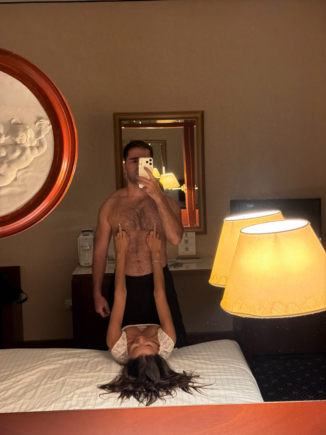A man and a woman are reflected in a mirror in what appears to be a hotel room. The man, partially undressed, is holding a smartphone and taking a selfie. The woman is lying upside down on a bed, facing the camera, with her arms raised and hands gesturing with middle fingers. The room has a large ornate mirror, a decorative wall sconce, a bed with white linens, and two lampshades visible. The overall lighting is dim. The location is not specified as Verona, Italy, nor can it be determined from the image.