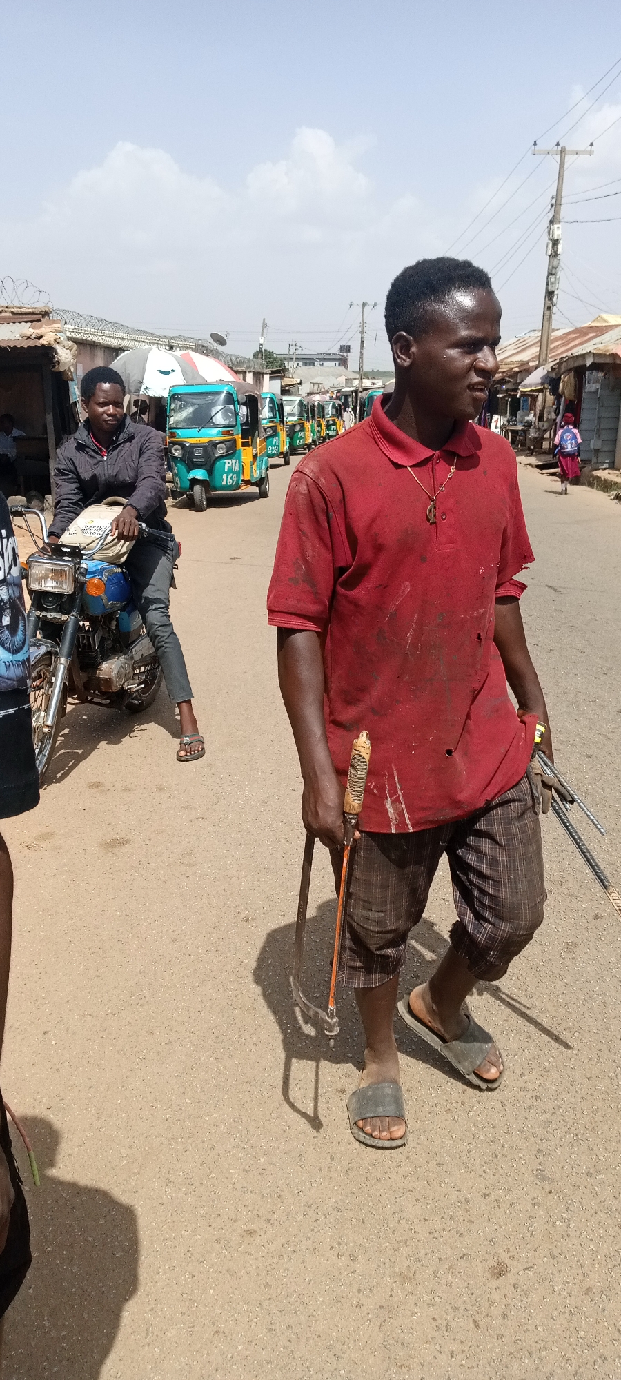 This image captures a bright, sunny scene of a bustling street in Jos, Nigeria, likely during midday due to the clear sky, scattered white clouds, and sharp shadows. The setting appears to be an urban or semi-urban commercial area, with various buildings, street vendors, and vehicles.
In the foreground, slightly to the right, a dark-skinned man walks, looking to his right. He wears a red polo shirt, visibly stained with white marks, plaid shorts, and grey slide sandals. A gold chain with a distinct pendant is visible around his neck. He carries tools associated with construction or metalwork: a hacksaw-like tool with a decorative, possibly carved, handle in his left hand, and several metal rods or pieces of rebar in his right. His expression is engaged, with a slight upward curve to his mouth.
On the left side of the midground, another dark-skinned man is seated on a blue motorcycle, holding the handlebars. He wears a dark jacket, dark trousers, and grey sandals, with a light-colored bag slung over his shoulder. He appears to be either riding slowly or momentarily stopped.
The street is made of dirt or a light pavement. In the background, a line of several colorful auto rickshaws (Keke Napep), predominantly green and blue, stretches down the road. One blue rickshaw in the middle ground clearly displays "PTA 169" on its front license plate area. Along the sides of the street, there are various low-rise buildings, some with corrugated iron roofs and some featuring barbed wire or razor wire fences. Utility poles with overhead wires are also visible. In the far distance on the right, a small child, possibly a girl, wearing a pink outfit, is walking away. The overall scene depicts typical daily activity on a vibrant Nigerian street.